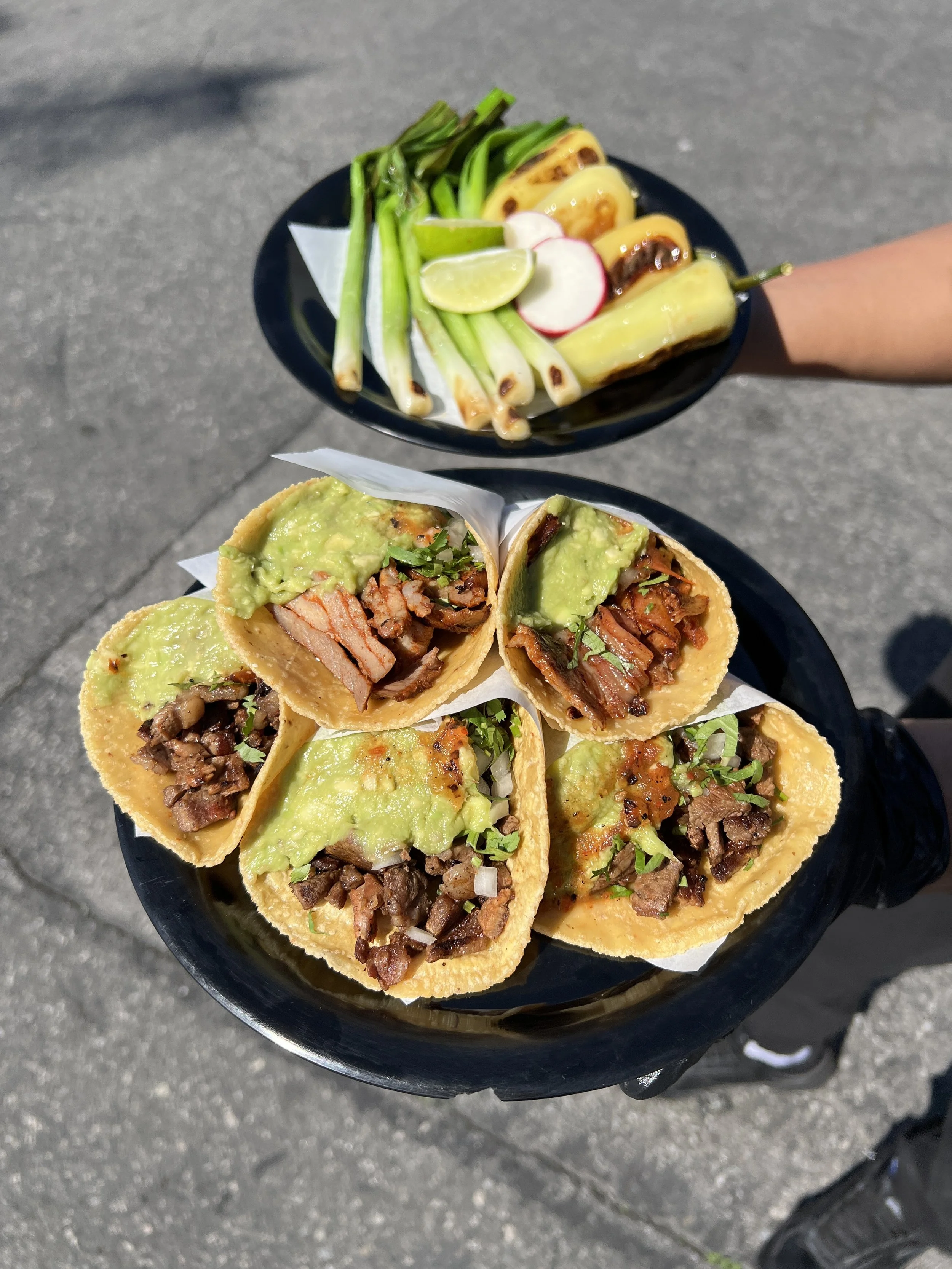 A person holding two black plates with Mexican food outdoors, featuring tacos filled with grilled meat, guacamole, and garnished with chopped onions and cilantro, and in the background, a plate of grilled green onions, radish slices, lemon wedges, and grilled jalapeños.