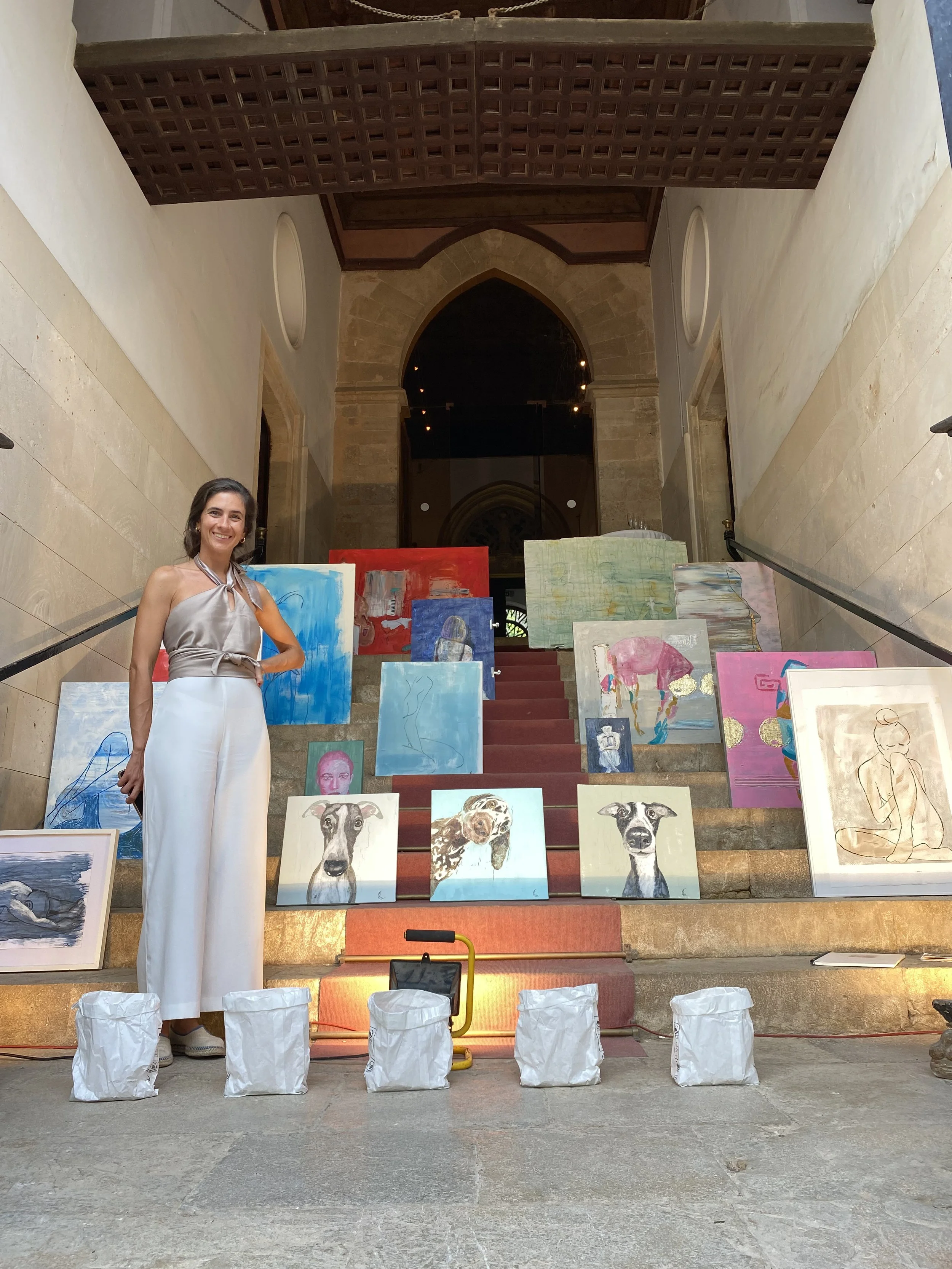 A woman standing on a staircase in an art gallery or museum, surrounded by colorful paintings, with some wrapped or covered objects in front of her. The background features an archway and large stone walls.