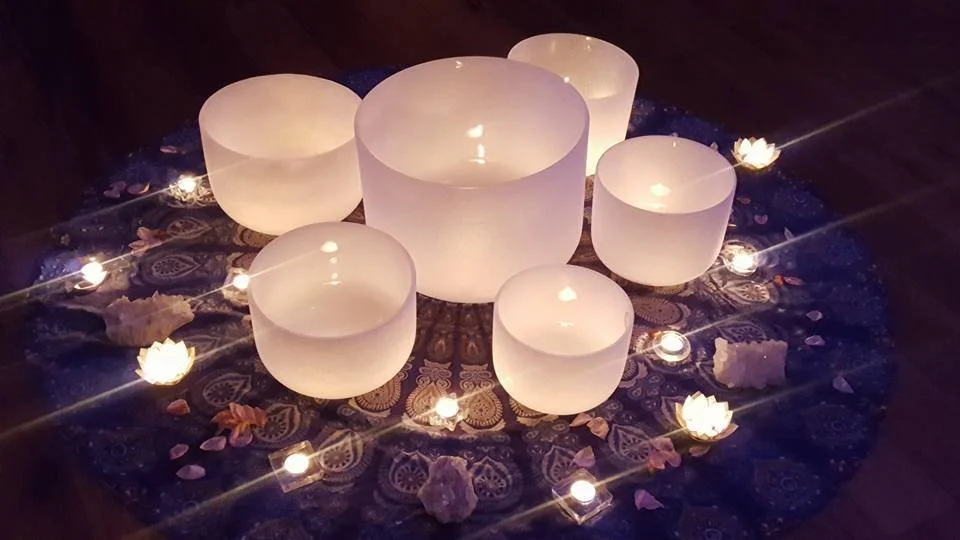 Seven white singing bowls of various sizes arranged in a circle on a decorative cloth, surrounded by small candles, rose petals, and flower-shaped lights, creating a relaxing or meditative setting.