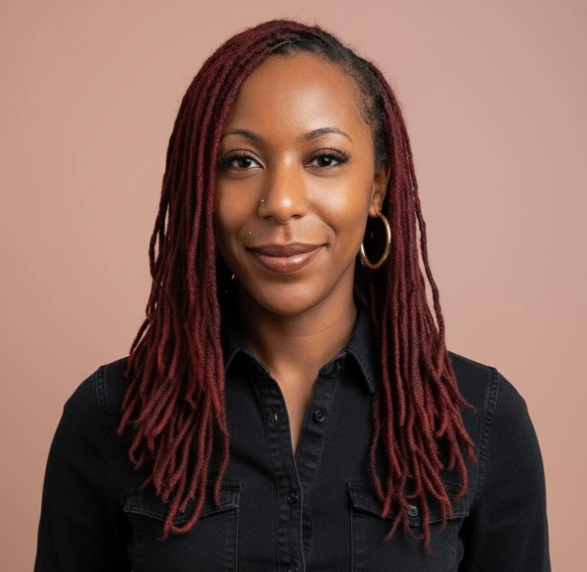A woman with long red dreadlocks, wearing hoop earrings and a dark button-up shirt, smiling in front of a beige background.