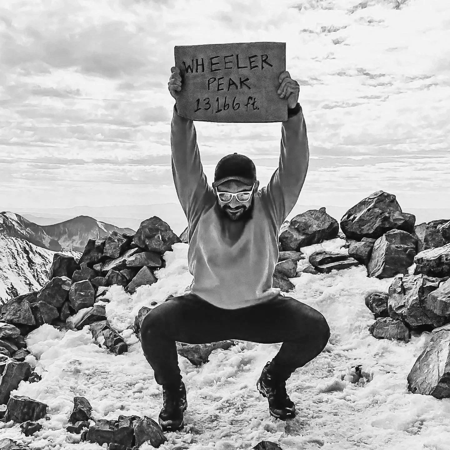 A person squatting on snow among rocks atop Wheeler Peak, holding a sign that reads "WHEELER PEAK 13166 ft."
