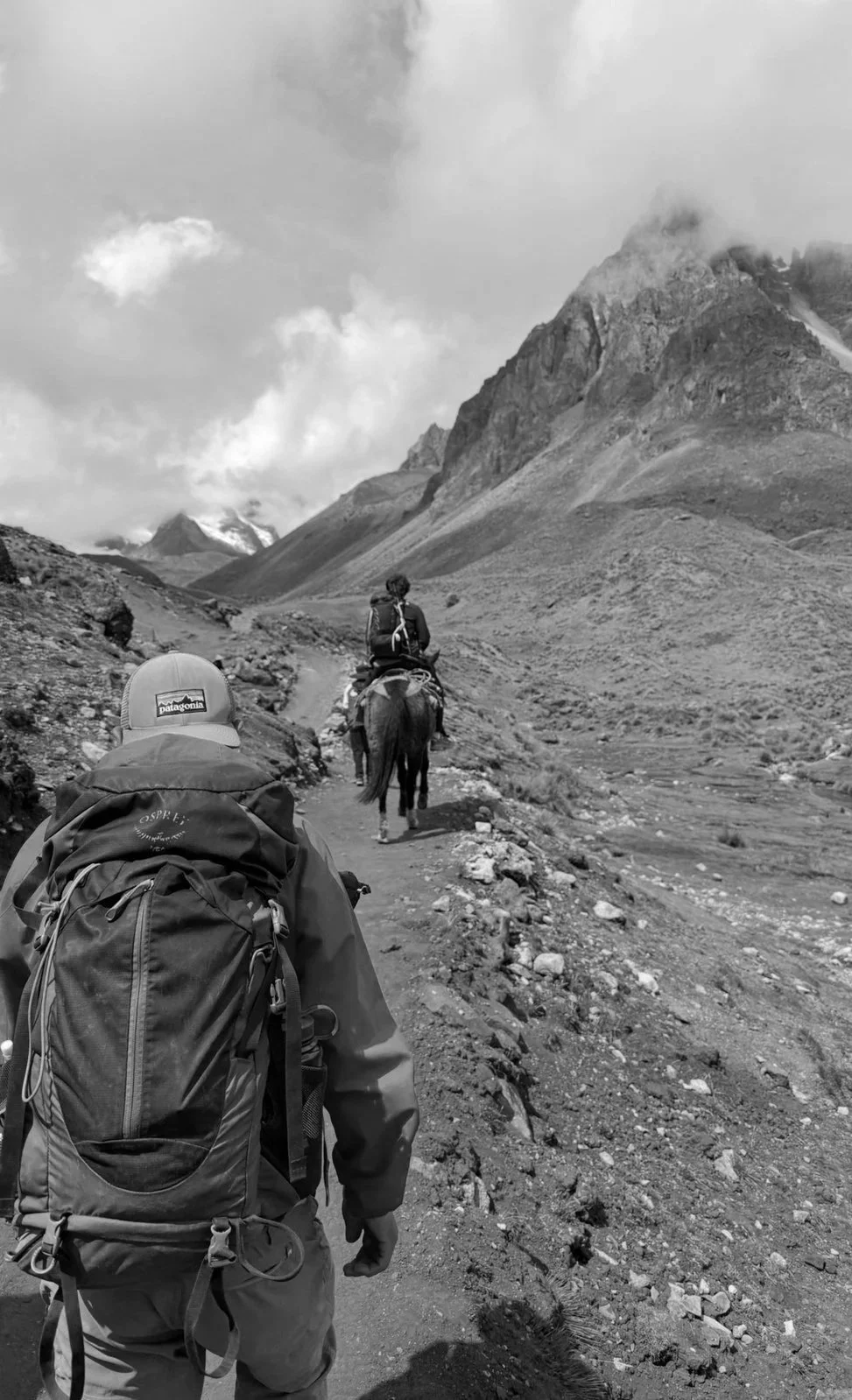 Hikers walking along a rocky mountain trail, one riding a horse, with mountains and cloudy sky in the background.