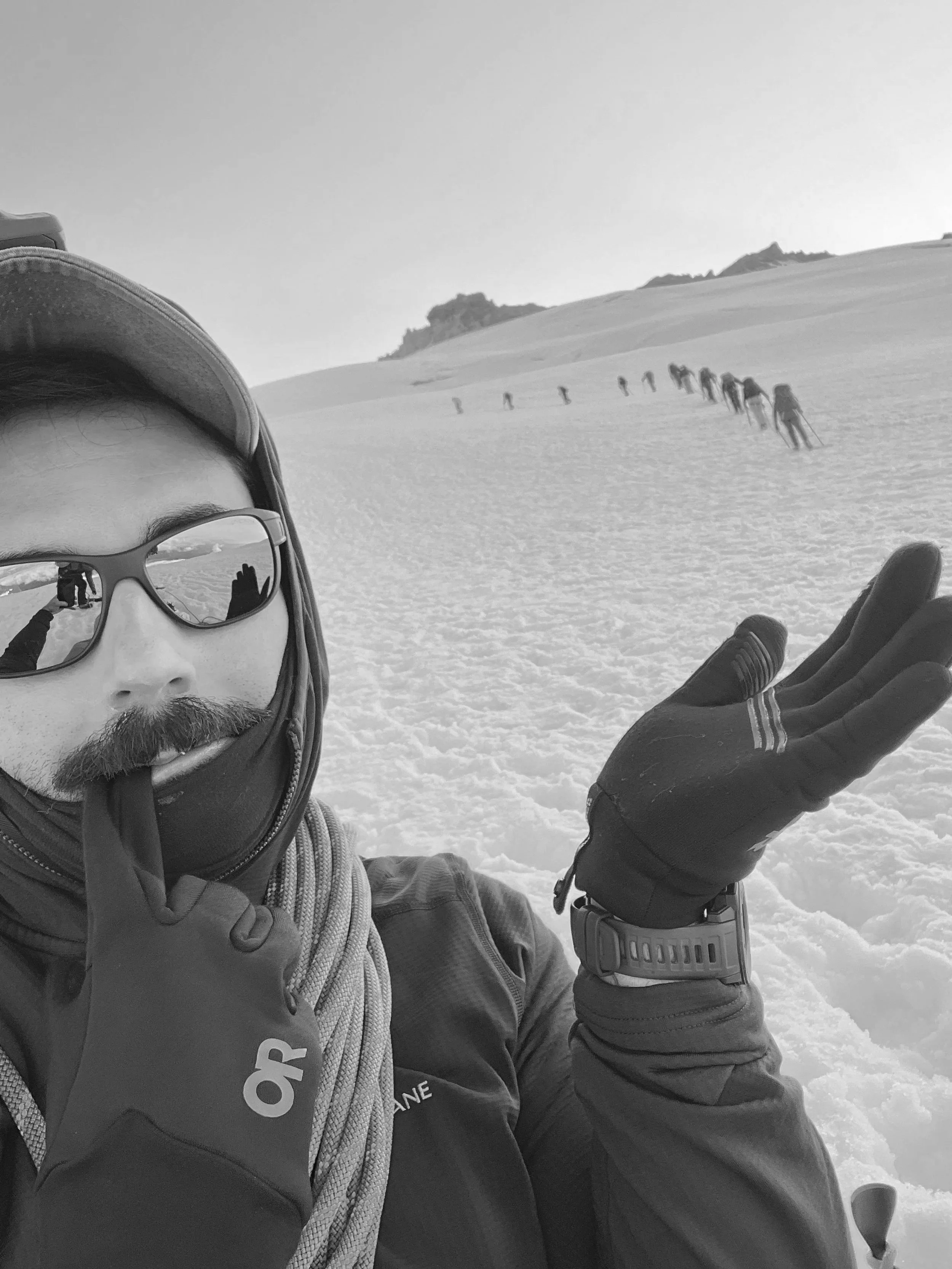 A person wearing sunglasses, a hat, and gloves taking a selfie on a snowy mountain with a line of climbers ascending in the background.