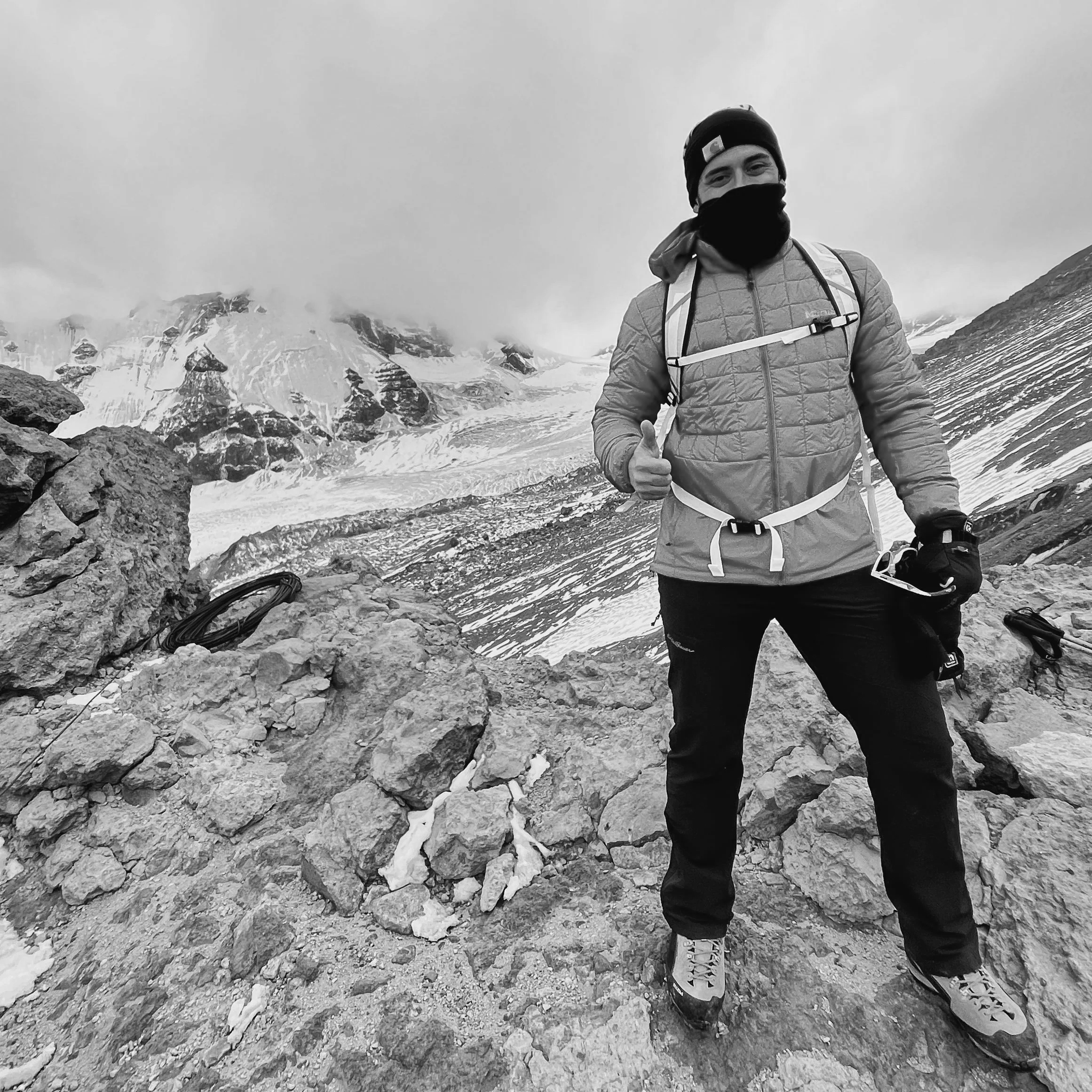 A man in outdoor winter gear standing on rocky terrain with a snow-covered mountain and glacier in the background.