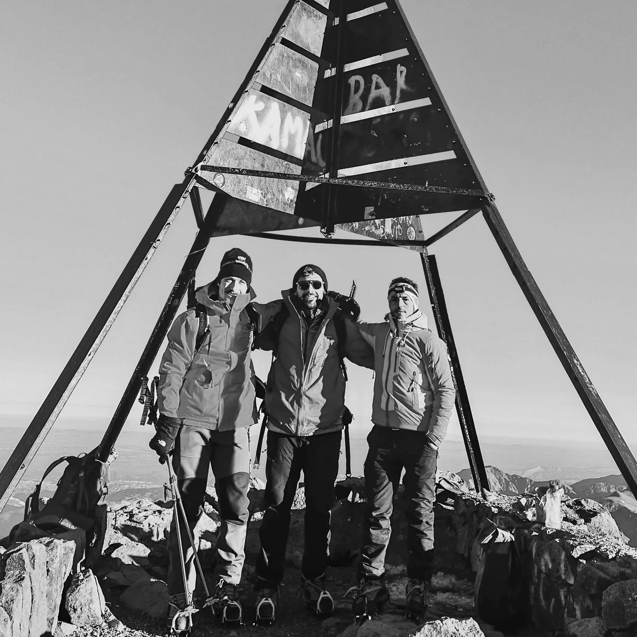 Three climbers standing together at a mountain summit under a triangular sign that reads 'RIM BAR', surrounded by rocks and distant mountains.