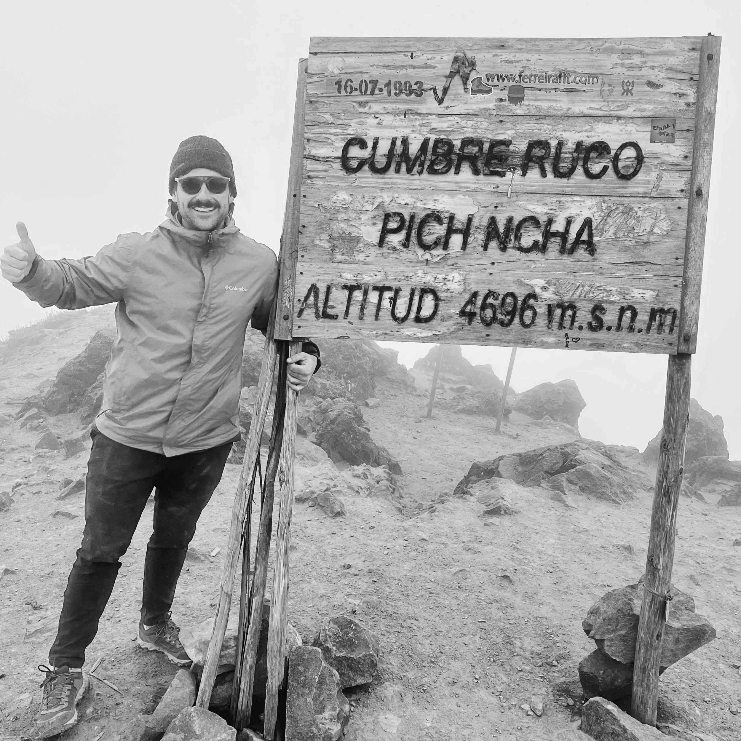 A man in outdoor clothing, wearing sunglasses and a beanie, standing next to a wooden sign on a mountain trail. The sign reads 'Cumbre Ruco Pich Ncha Altitud 4696 m.s.n.m.' The background is foggy with rocks and uneven terrain.