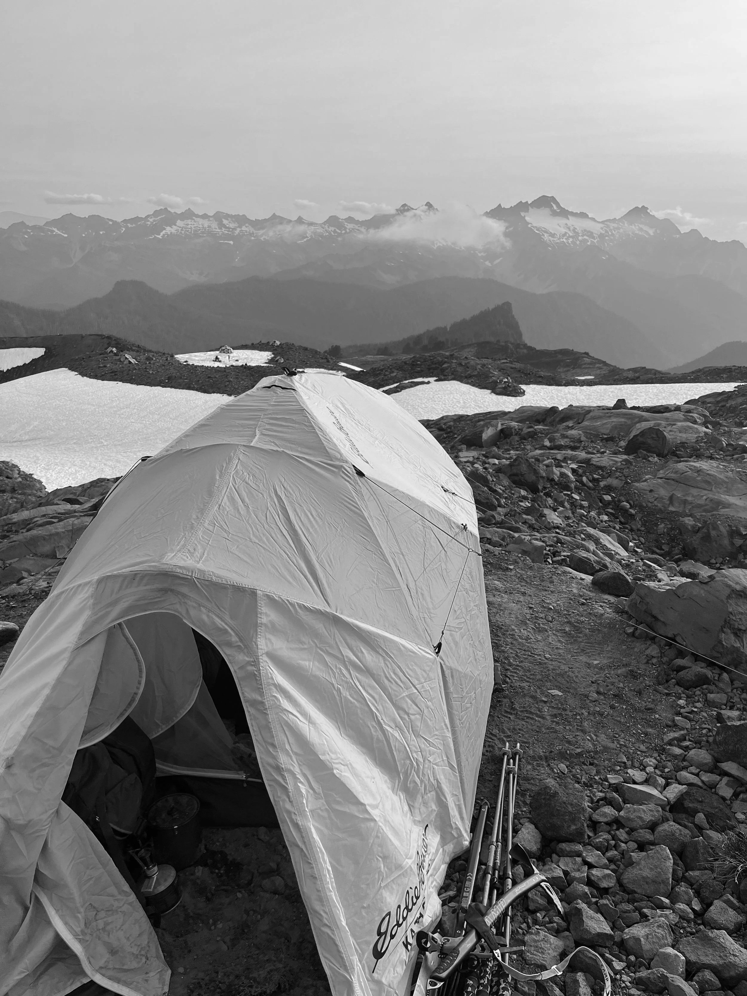 A white tent set up on rocky terrain in a mountainous landscape with snow patches, distant mountain peaks, and clouds in the background, in black and white.