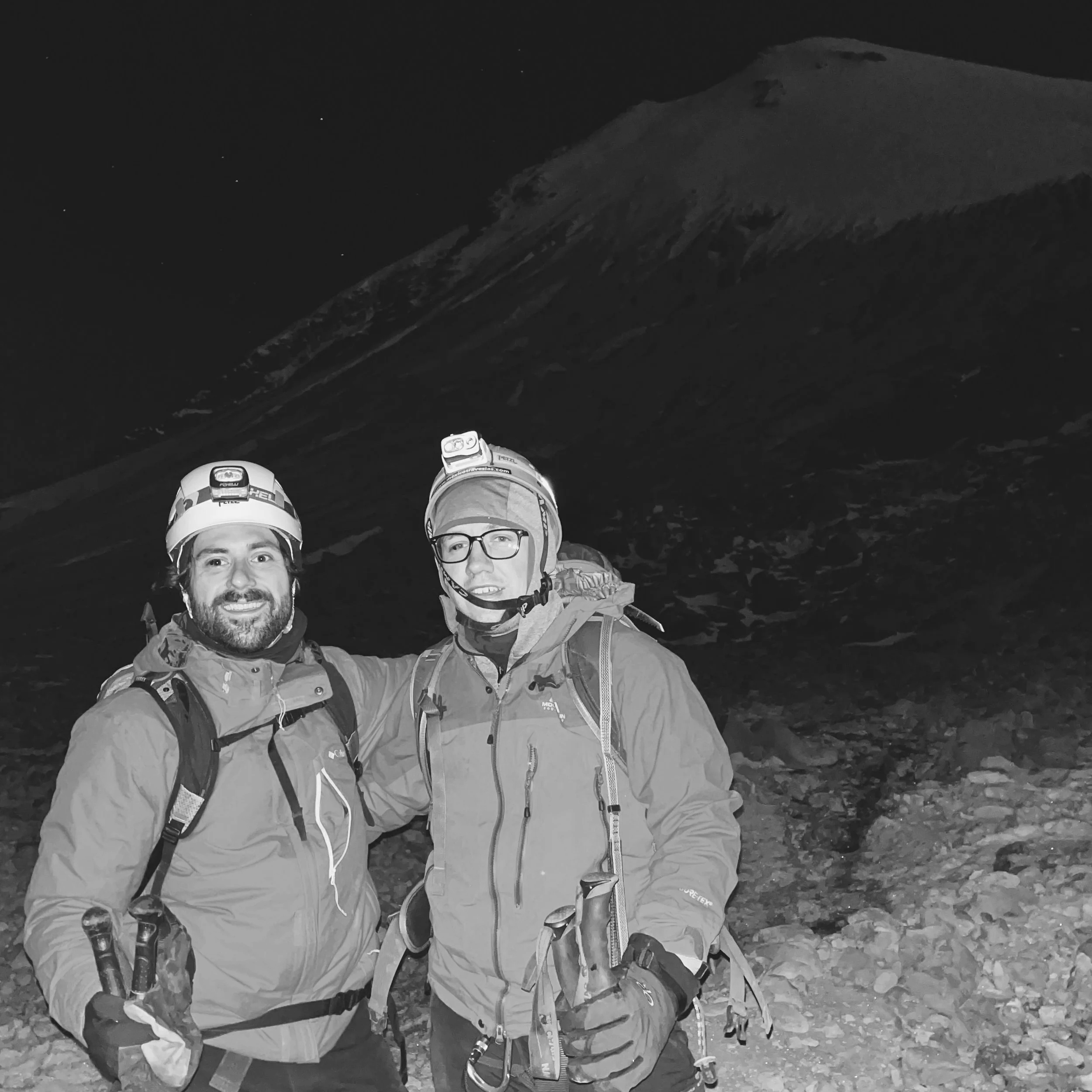 Two male climbers in outdoor gear, helmets, and headlamps, standing together at night with a mountain in the background.