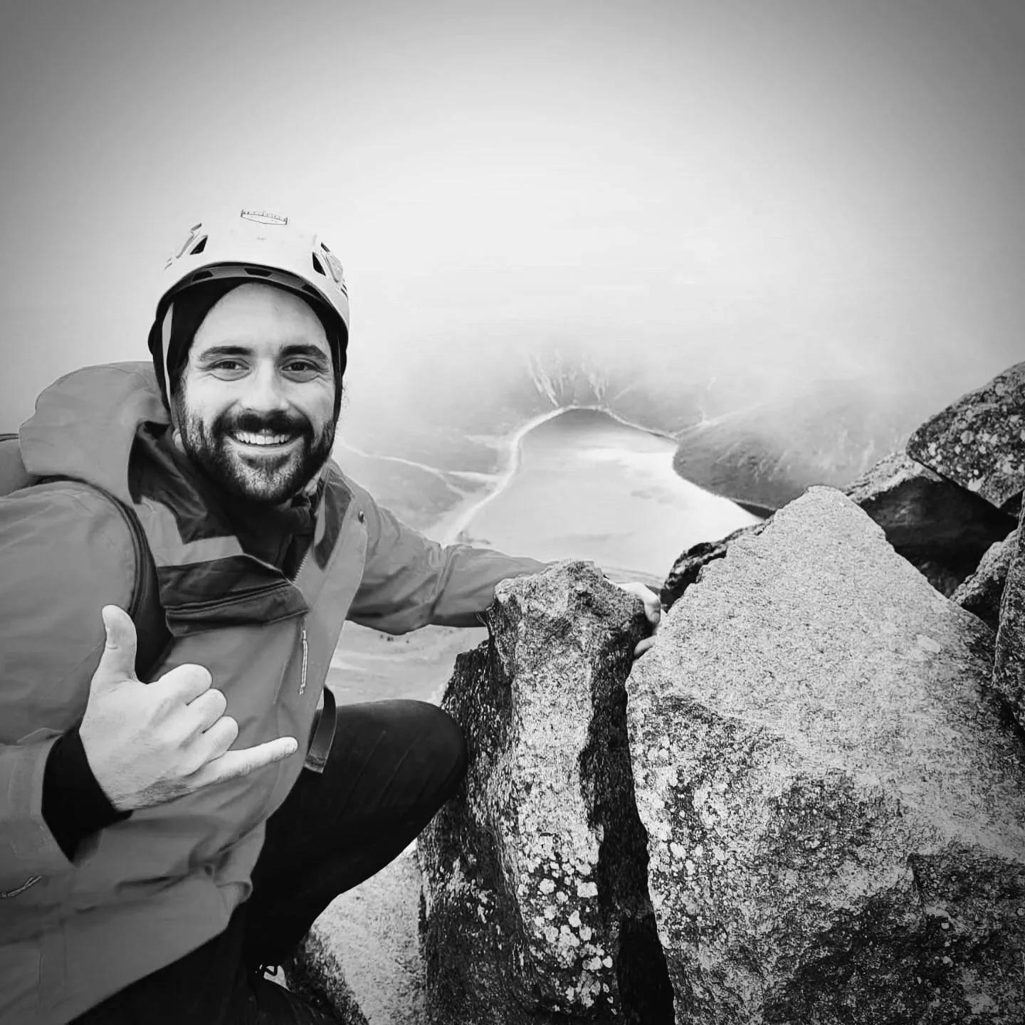 A man at a mountain summit taking a selfie near large rocks with a foggy landscape and a body of water in the background. The man is smiling, wearing a helmet, jacket, and giving a thumbs-up gesture.