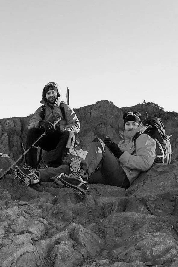 Two hikers in outdoor gear resting on rocky terrain with mountains in the background, one sitting and the other kneeling.
