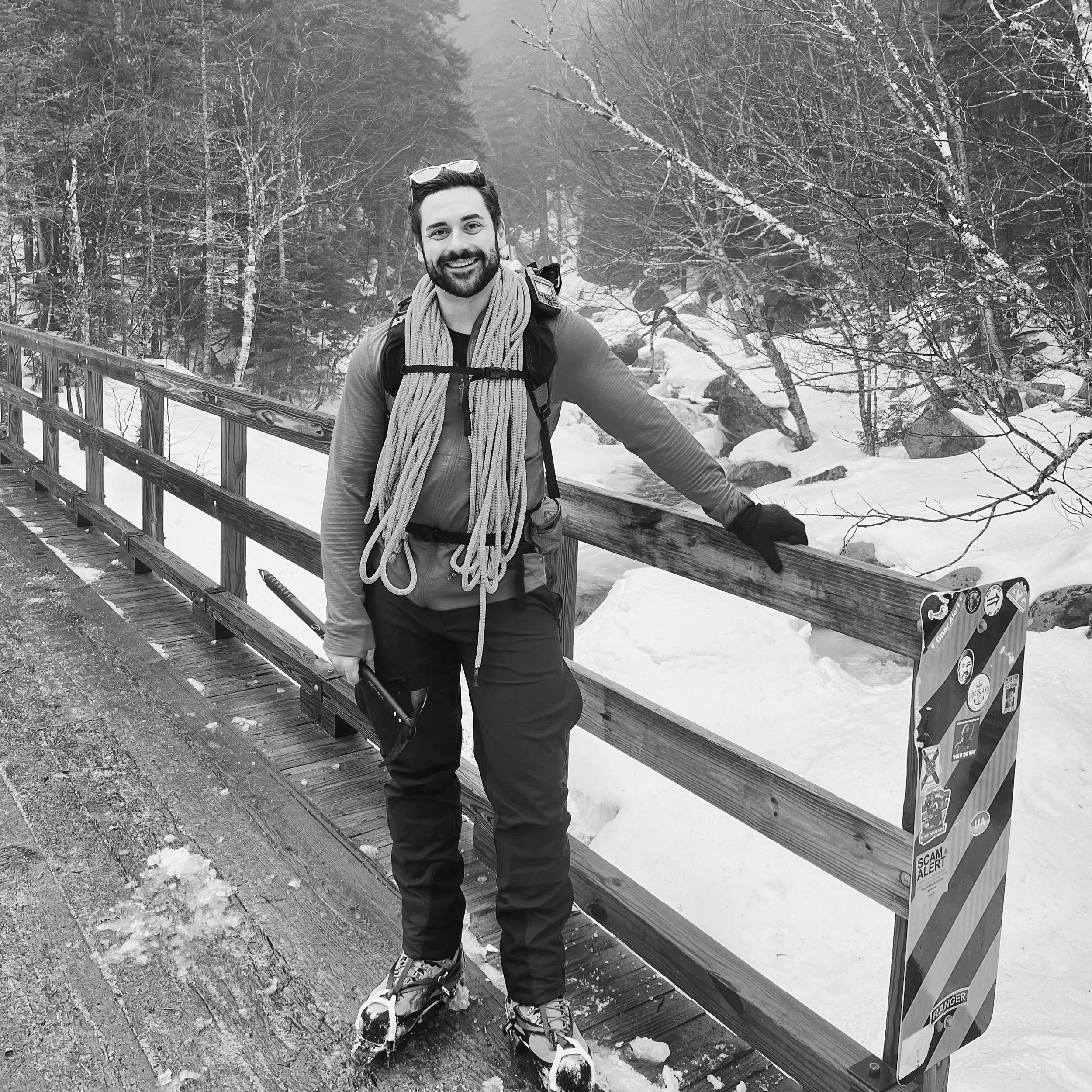 A man with a beard and smiling face, wearing outdoor gear including skis, stands on a snowy mountain road, leaning on a wooden fence in a winter forest.