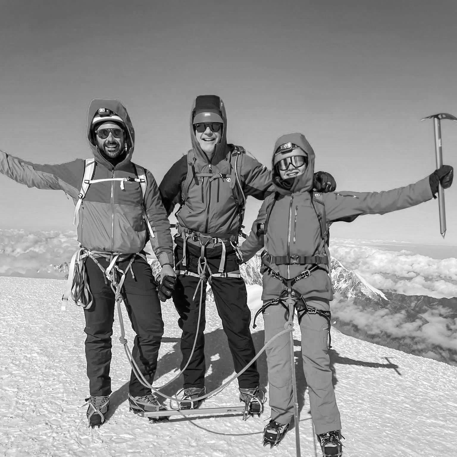 Three climbers in gear standing on snow-covered mountain summit with cloud and mountain landscape in background.