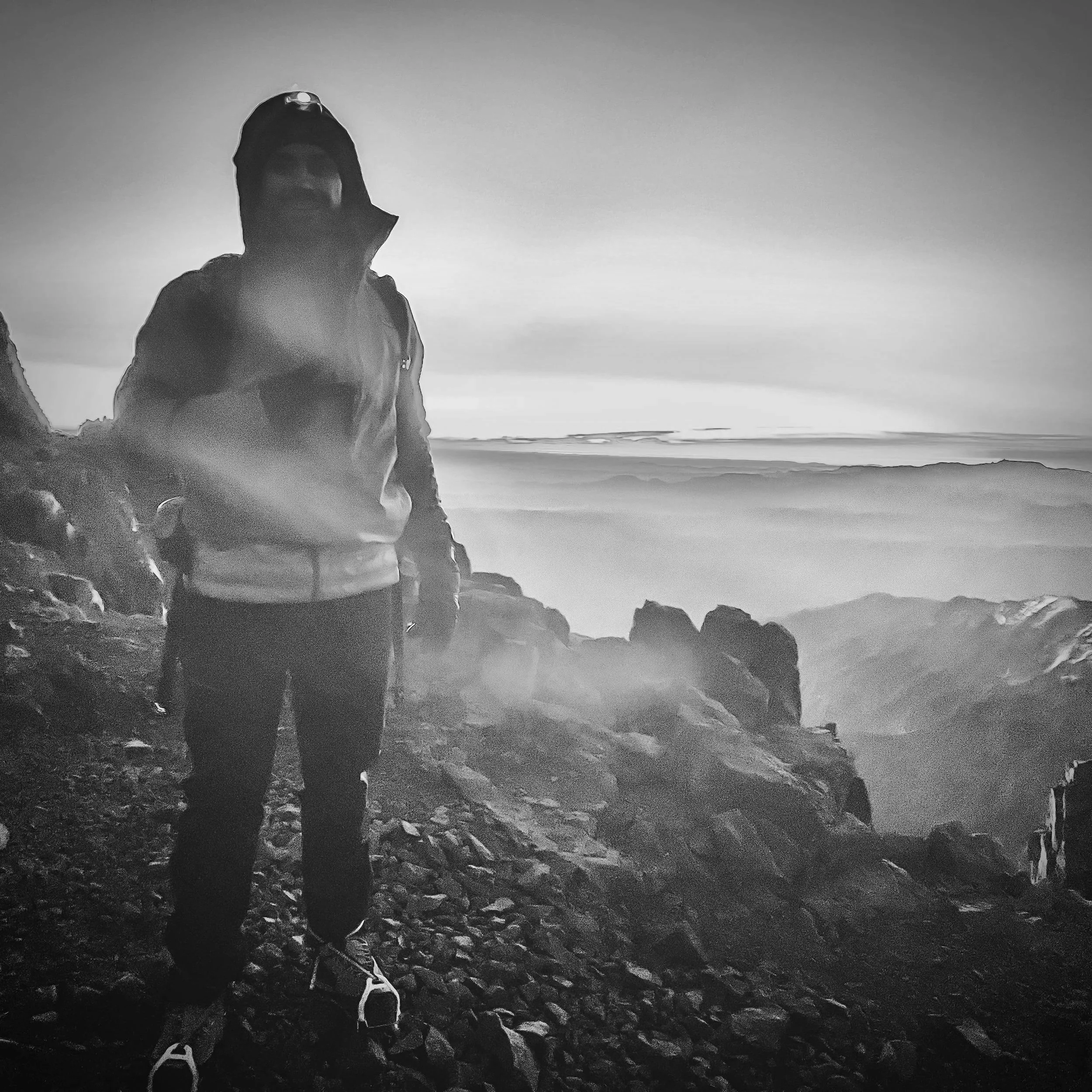 Person dressed in outdoor gear standing on rocky terrain near a cliff with misty mountains and sky in the background, black and white photo.