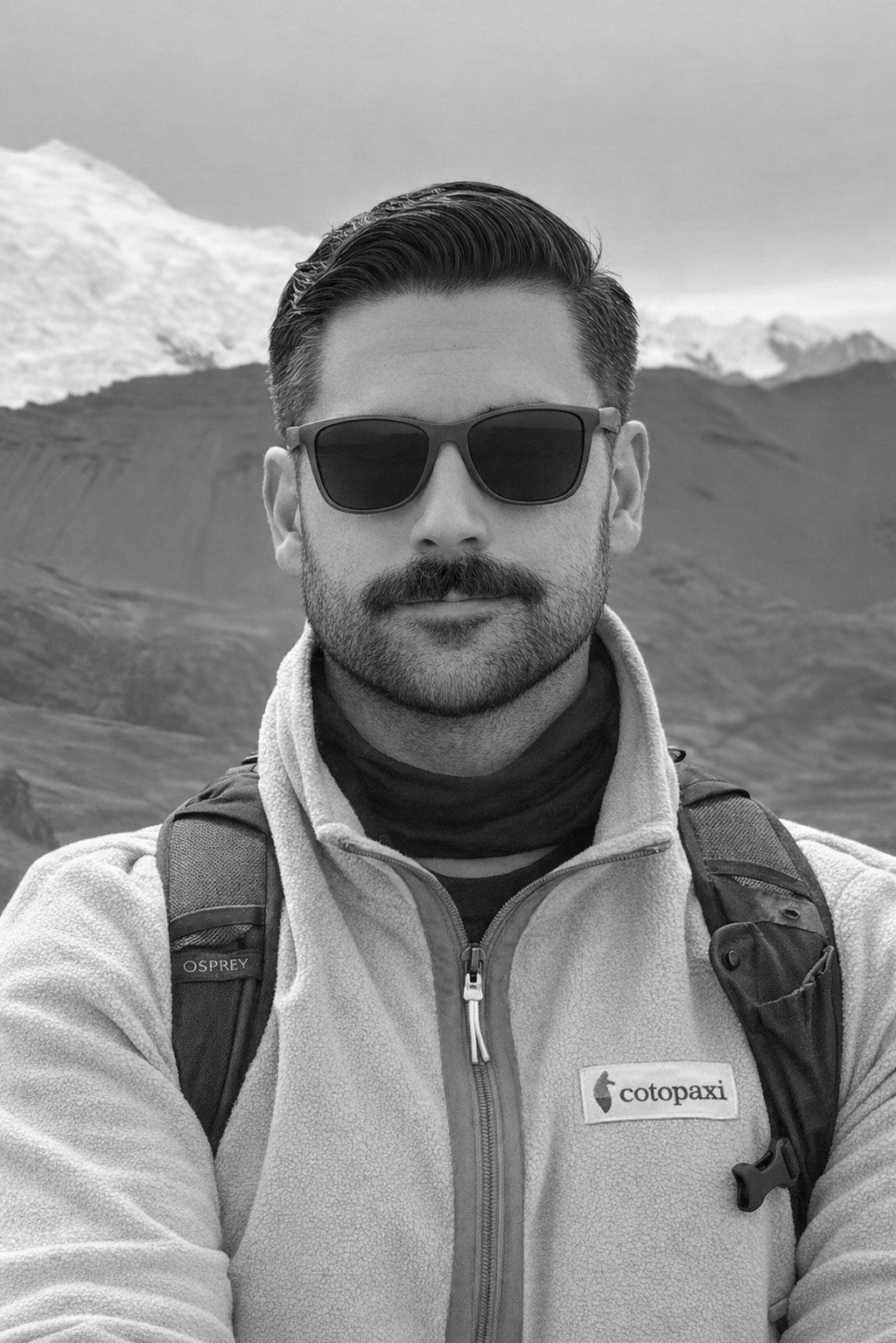 A man wearing sunglasses, a fleece jacket with a zip, and a backpack taking a selfie outdoors with snowy mountains in the background.