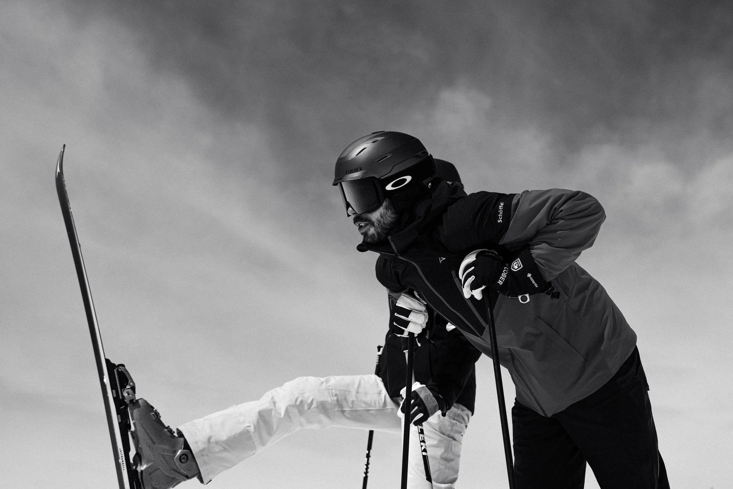 Two skiers, one in a black jacket and the other in a light-colored jacket, are leaning forward, one with a ski pole inhand. They are dressed in winter sports gear, including helmets and goggles, and seem to be on a snowy slope.