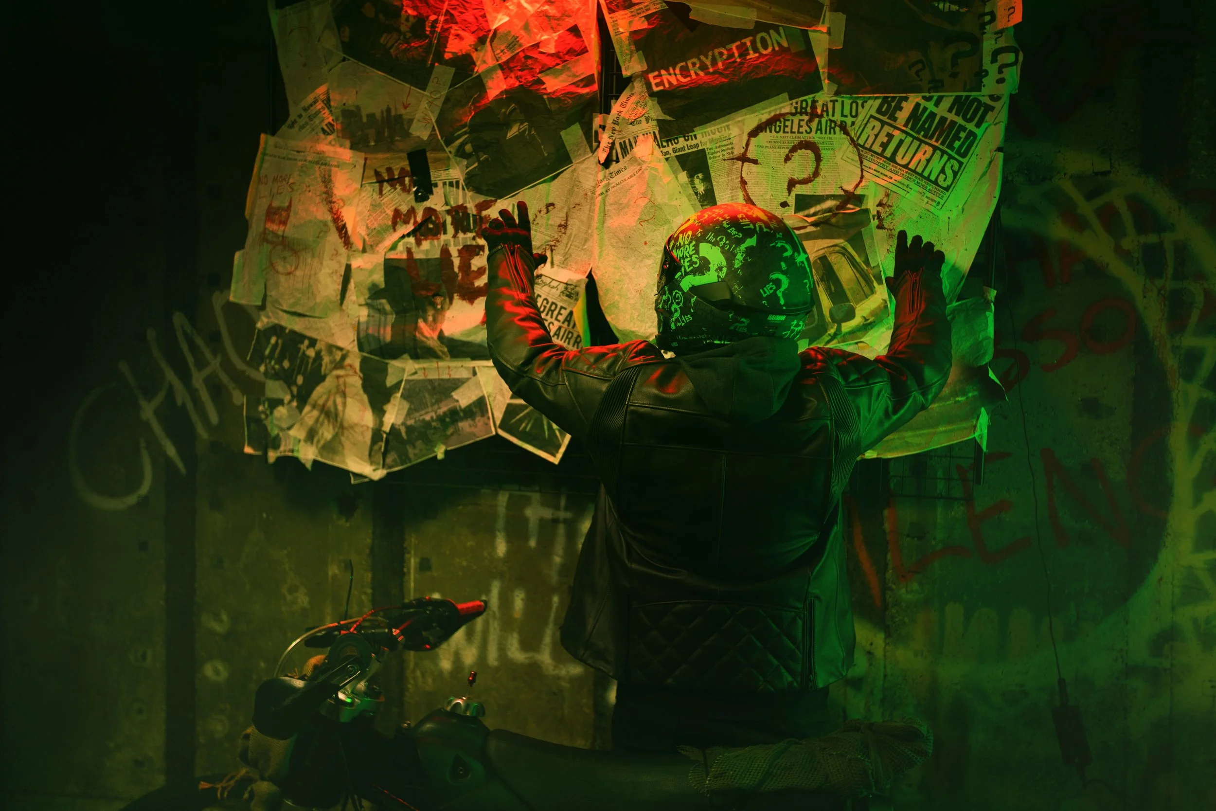 A person wearing a helmet and leather jacket, facing a wall covered with newspapers, graffiti, and signs, with their arms outstretched, appearing to tag or draw on the wall in a dimly lit environment illuminated by green and red lights.