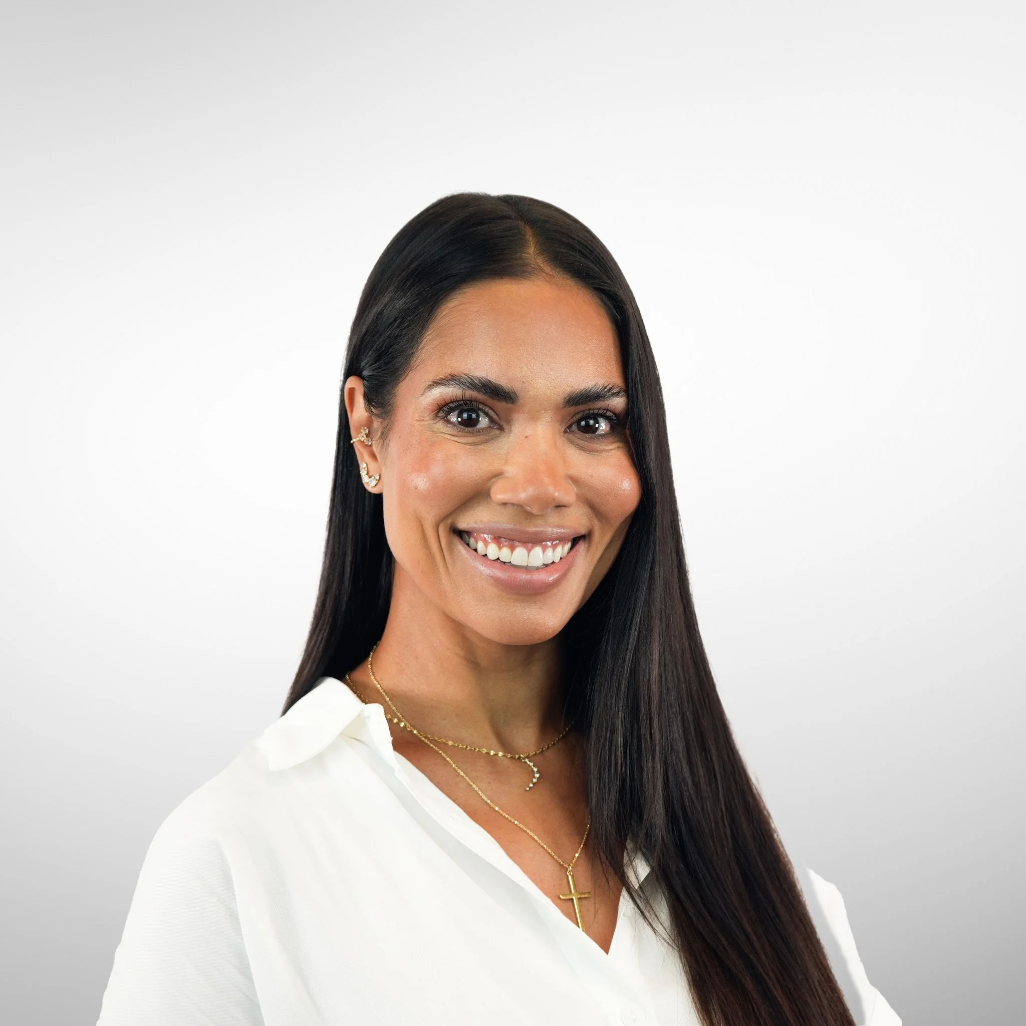 Smiling woman with long dark hair wearing a white shirt and gold jewelry, against a light gray background.