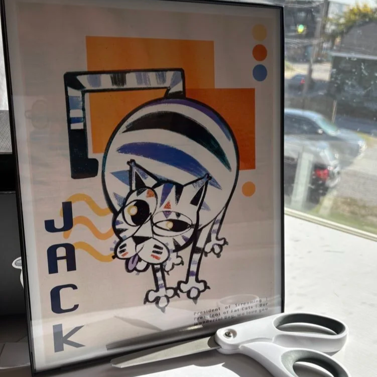 A colorful sign featuring a cartoon cat with sunglasses, standing in front of a striped umbrella. The sign has the word 'JACK' written vertically on the left side. There are abstract shapes and colors in the background, and a pair of scissors is placed in front of the sign.