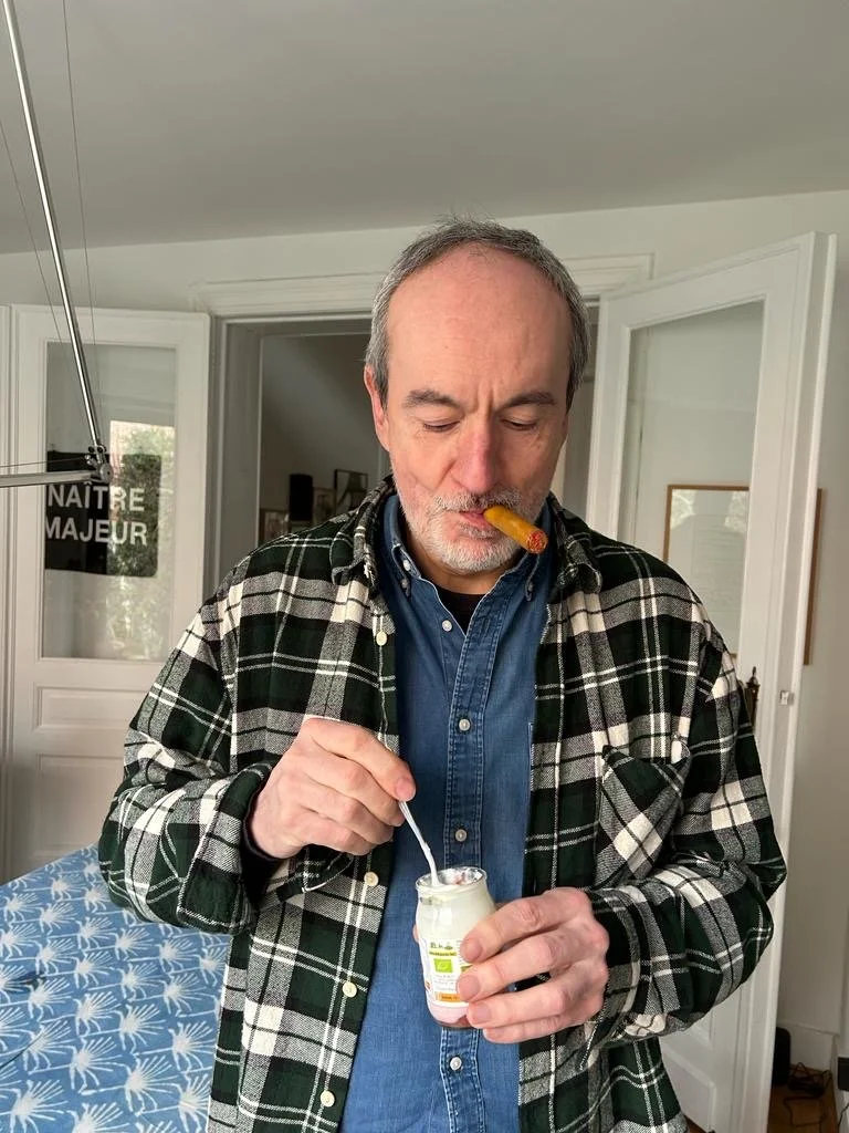 A man with gray hair and a beard, wearing a blue shirt and plaid jacket, stands indoors holding a small container of yogurt with a spoon, with a cigar in his mouth and a playful expression.