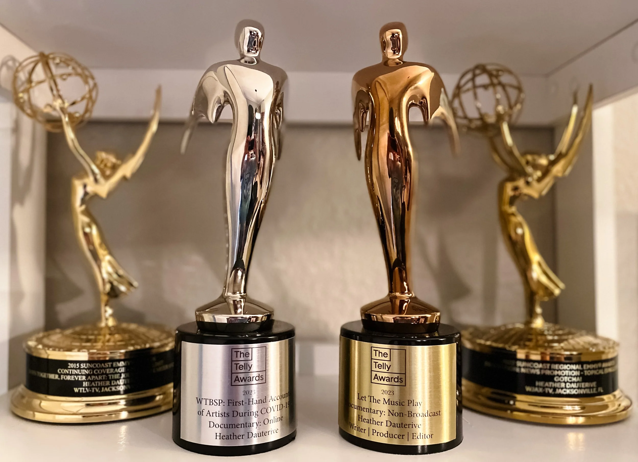 Three awards trophies with black and gold bases, featuring a female figure with outstretched arms as the main design, displayed on a white shelf with other spherical decorative objects in the background.