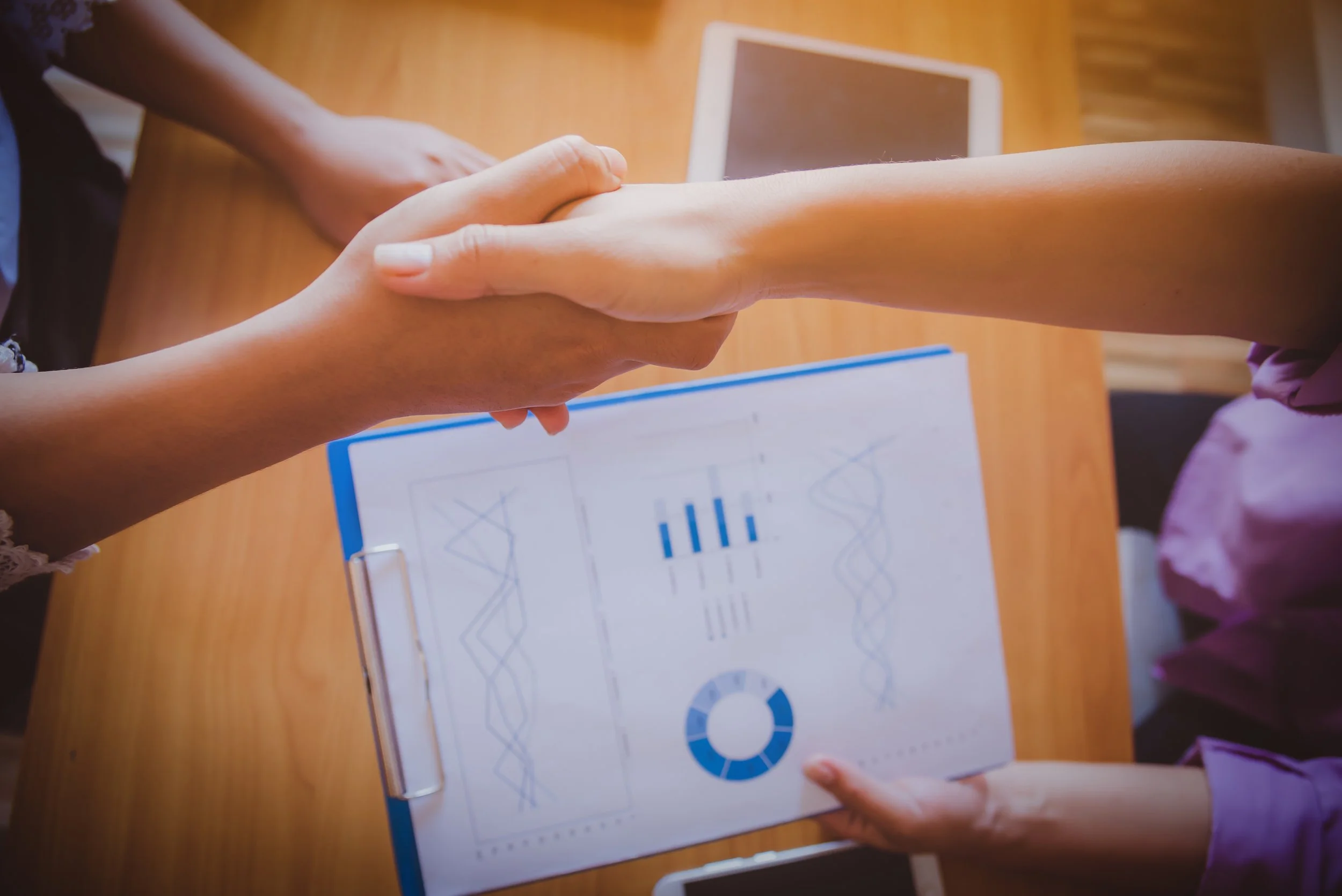Hands holding a sheet of paper with various scientific graphs and diagrams, with a tablet and a clipboard with more graphs on a wooden table.