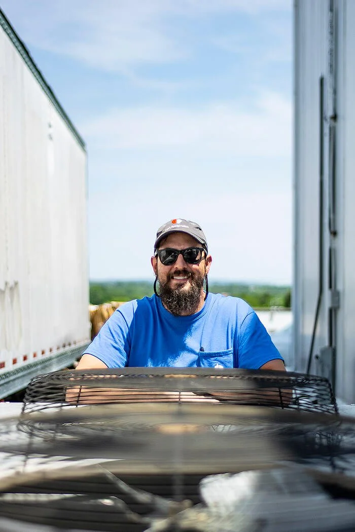 Smiling HVAC repair professional man with beard, sunglasses, grey cap, and blue t-shirt, standing between two semi-truck trailers outdoors on a sunny day.