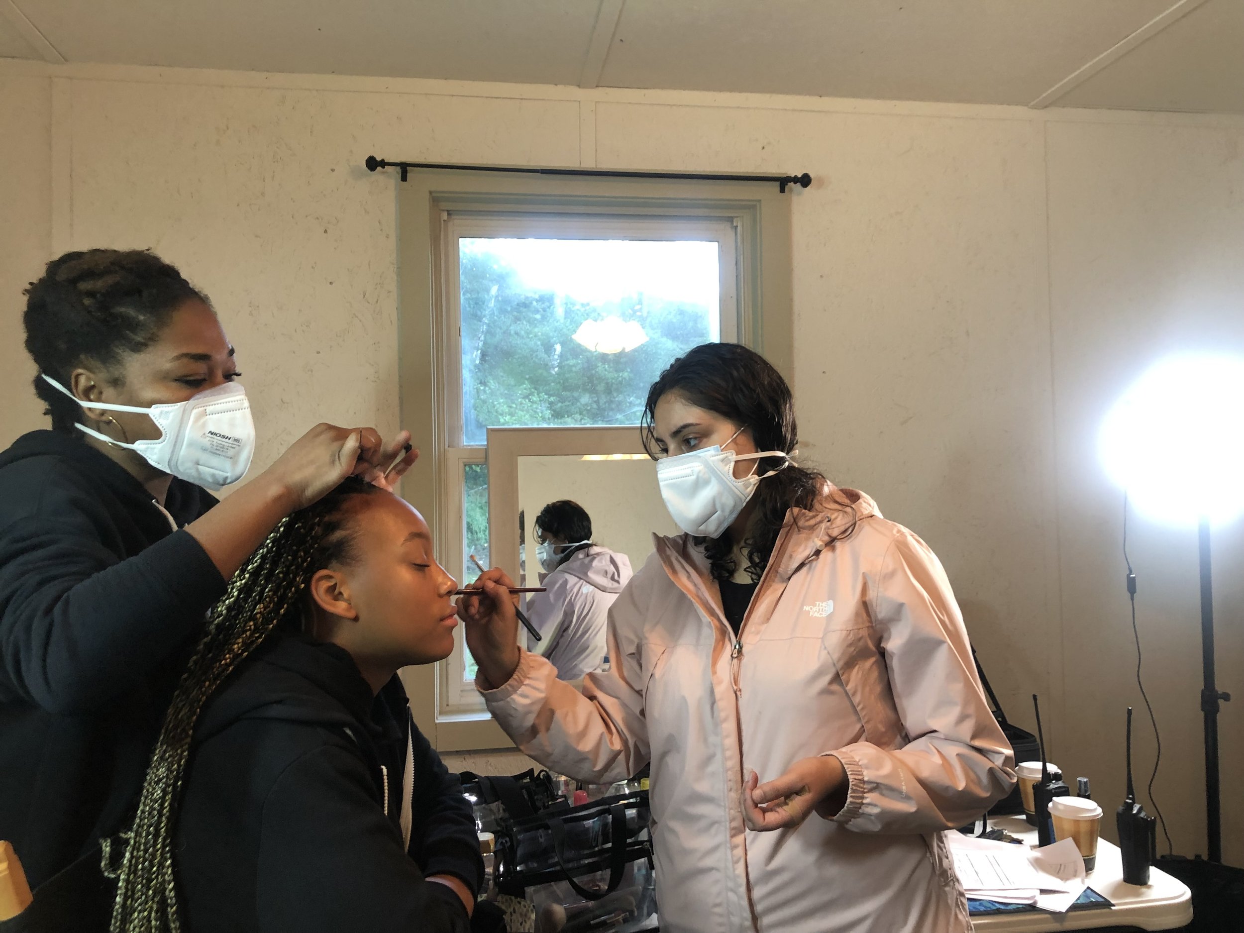 Makeup artists applying full glam makeup on a young woman's face in a room with a window and bright light.