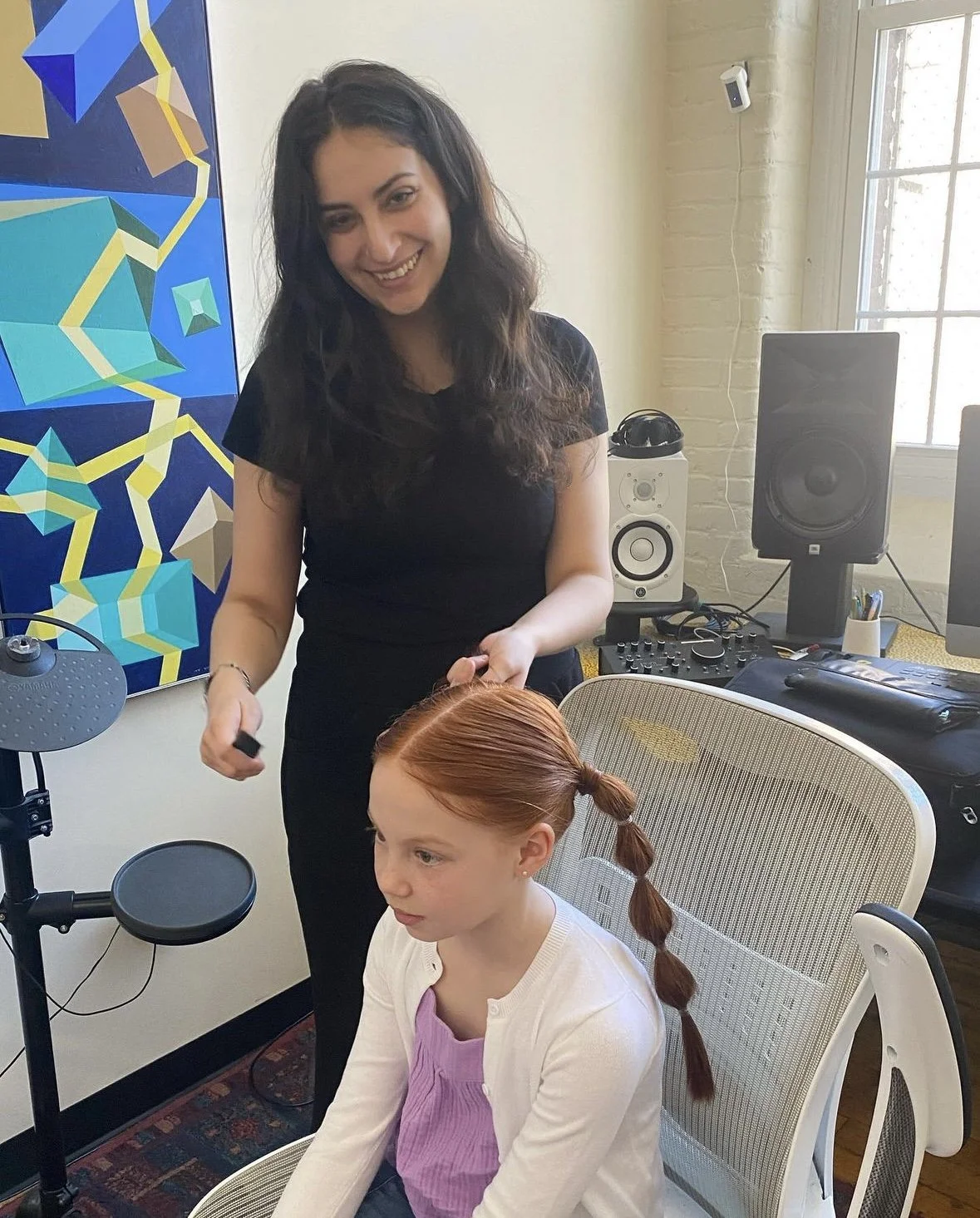 A woman with dark, wavy hair is styling a young girl with red hair, who is sitting in a chair. The woman is smiling and holding a hairbrush, while the girl has her hair styled in a braid. The room has professional audio equipment, including speakers, a mixer, and computers, with a colorful geometric painting on the wall.