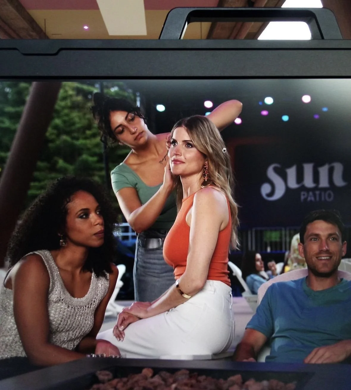 Group of four people sitting at a canopy-covered outdoor patio at sunset, with one woman receiving makeup or touch-up from makeup stylist