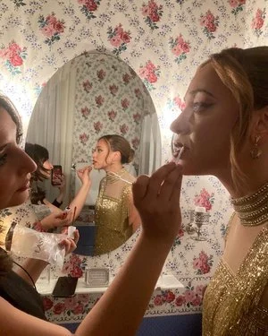 A woman with makeup and jewelry is getting her makeup done by a makeup artist in front of a mirror with floral wallpaper.