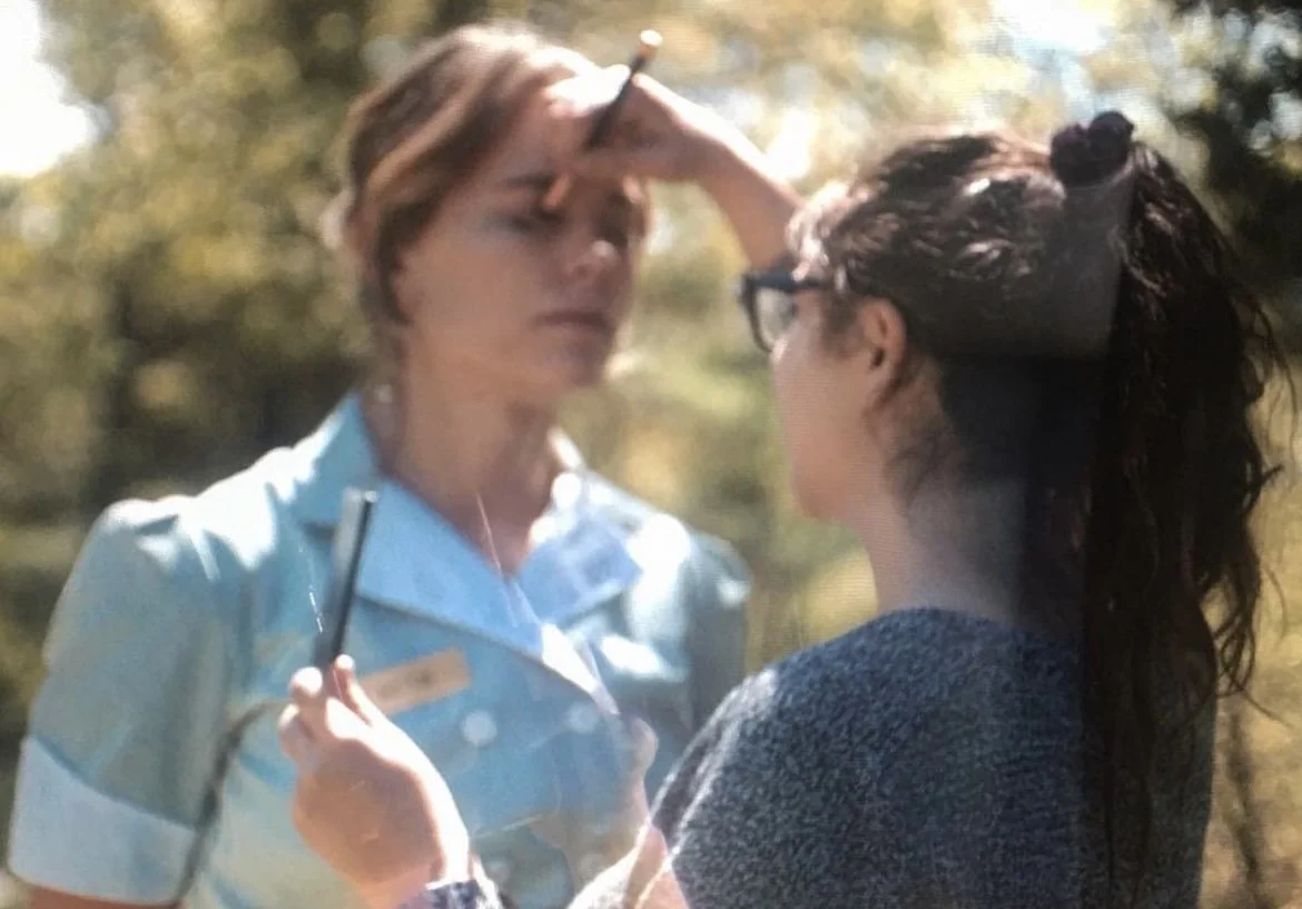 A woman in a light blue uniform is standing outdoors with her hand on her forehead, while another woman with dark wavy hair, glasses, and a dark sweater looks at her closely during daytime.
