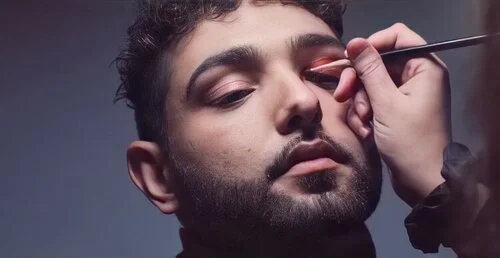 A man is getting his makeup done with a brush on his eyelid by a makeup artist.