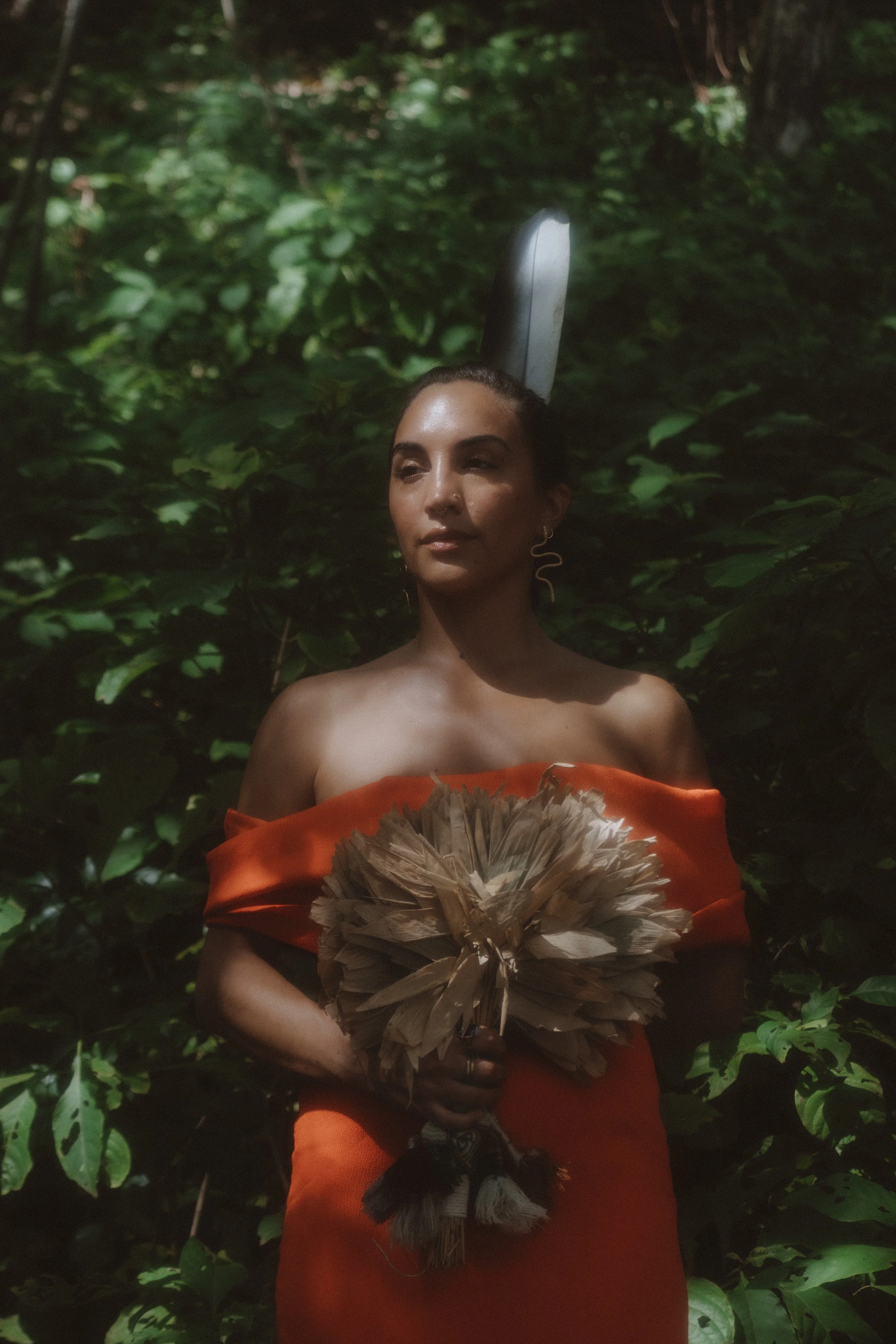 A woman standing outdoors among green foliage, holding a fan made of feathers, wearing an off-shoulder orange dress, with a dark, elongated object behind her head.