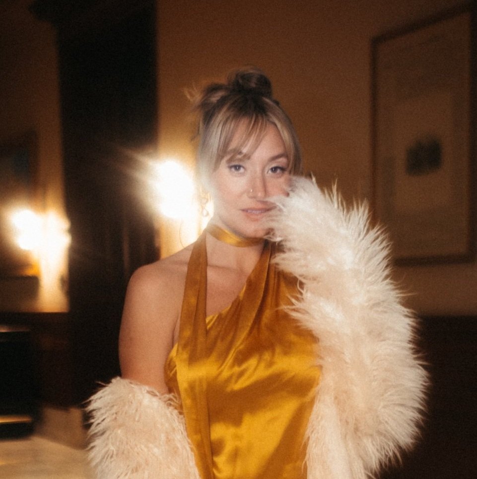 A woman in a gold satin dress and white fur coat, with her hair styled in an updo, standing indoors with backlighting and warm lighting fixtures.