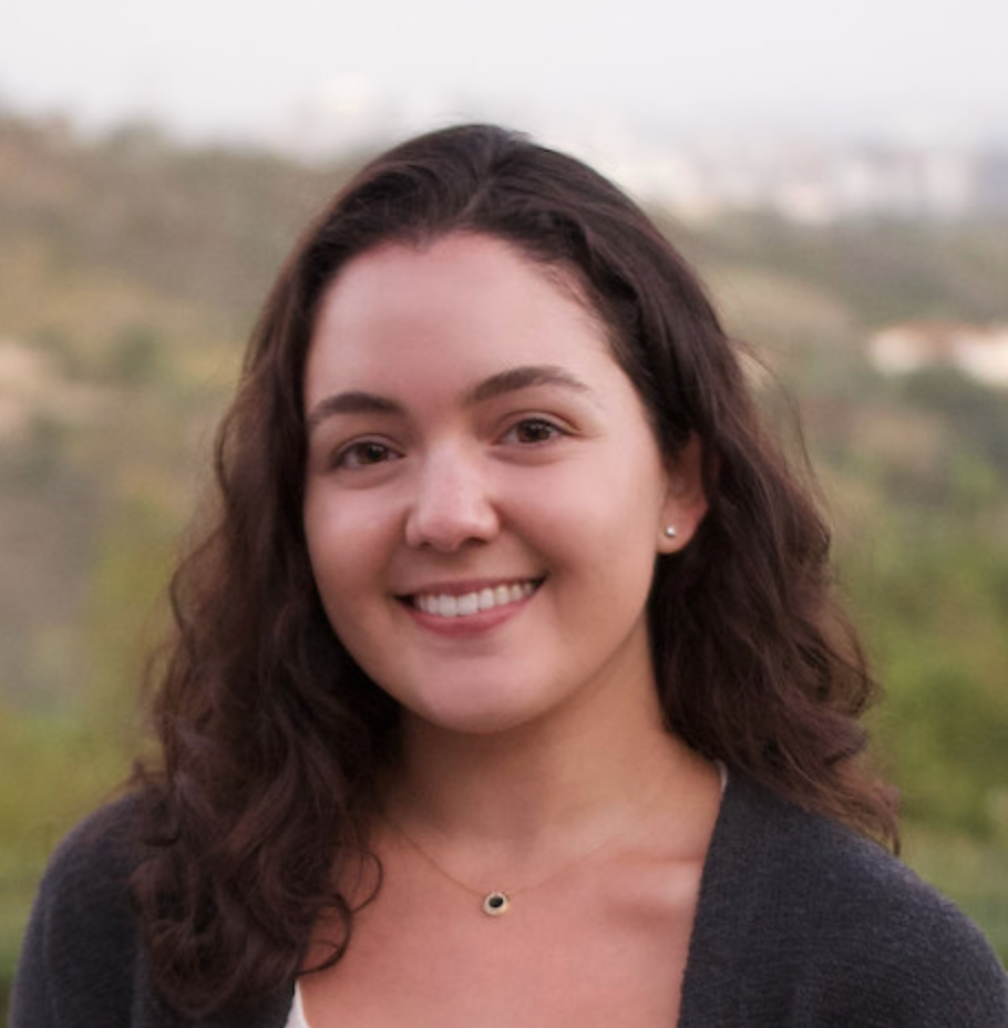 Close-up of a young woman with shoulder-length dark wavy hair smiling outdoors with a blurred background of trees and hills.