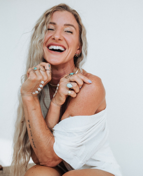 Young woman with long blonde hair smiling widely, wearing multiple rings and bracelets, holding her hands close to her face, dressed in a white off-shoulder top, against a plain light background.