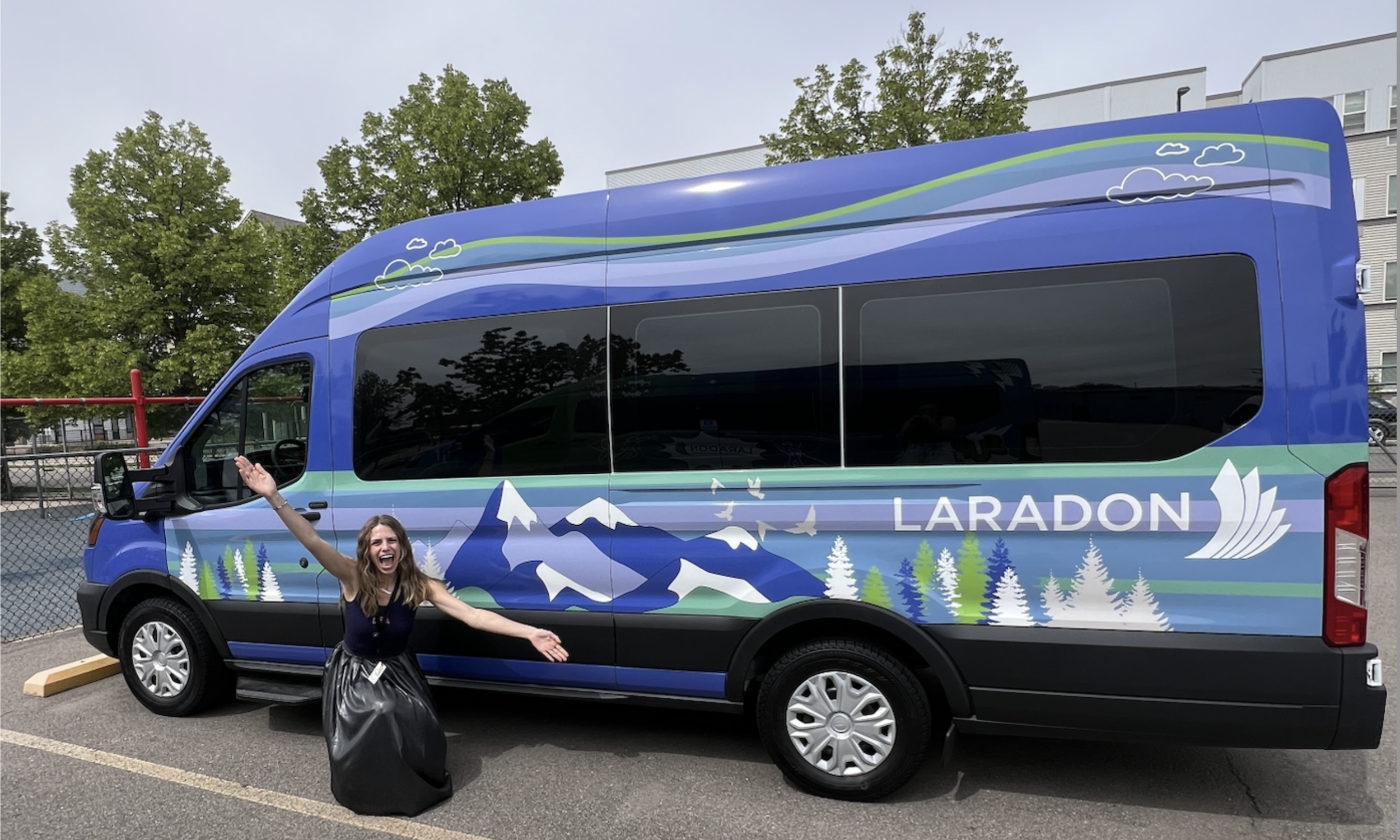 A woman standing joyfully with her arms outstretched in front of a blue Laradon Ford E-Transit electric van decorated with custom mountain and pine tree graphics.