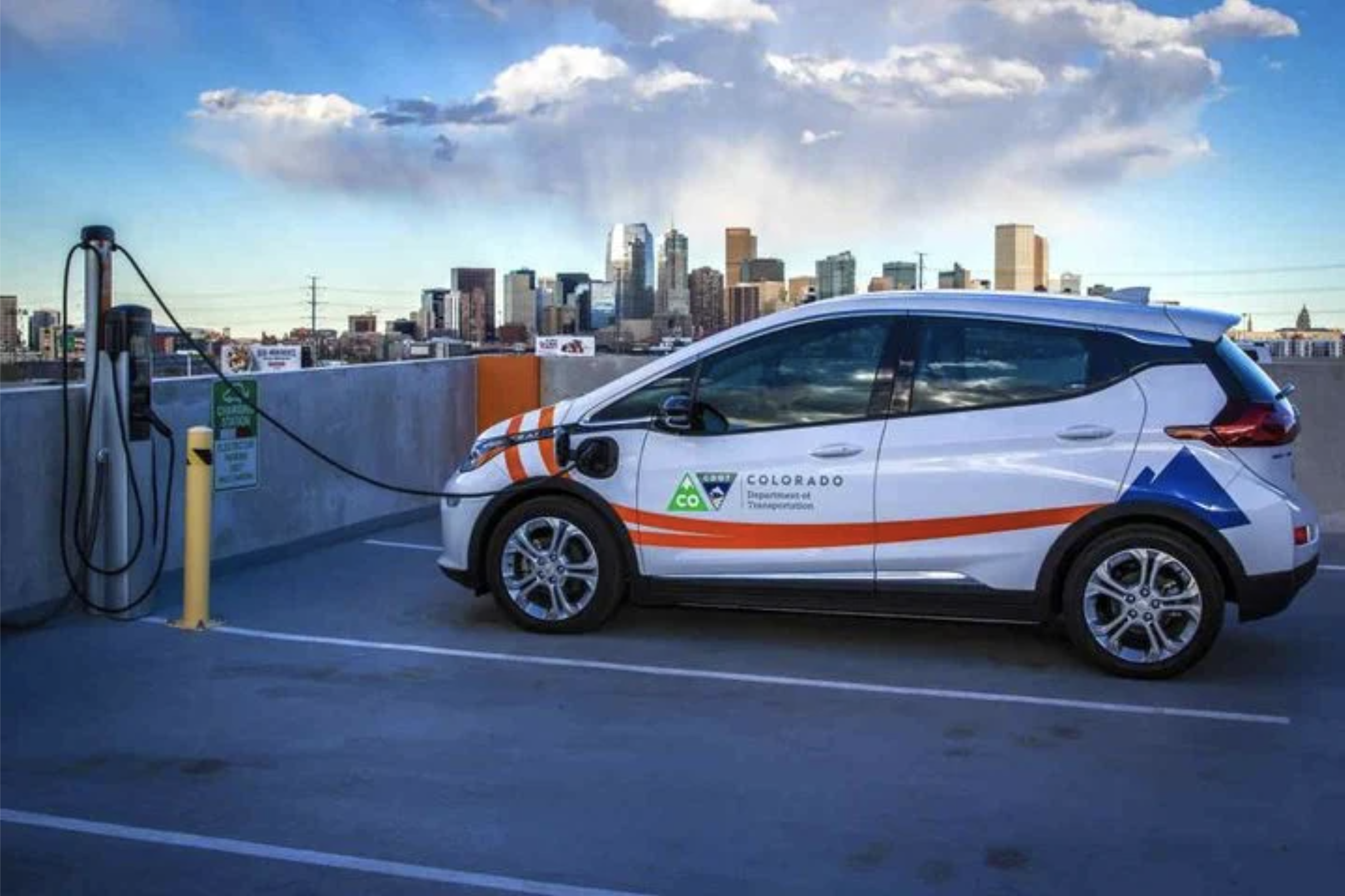 A white Chevrolet Bolt EV branded with Colorado Department of Transportation logos plugged into a charging station on a rooftop garage with the Denver skyline in the background.