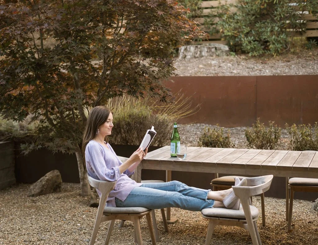 Woman sitting outdoors at a table, reading a book, with a bottle of water and a glass on the table, surrounded by trees and outdoor furniture.