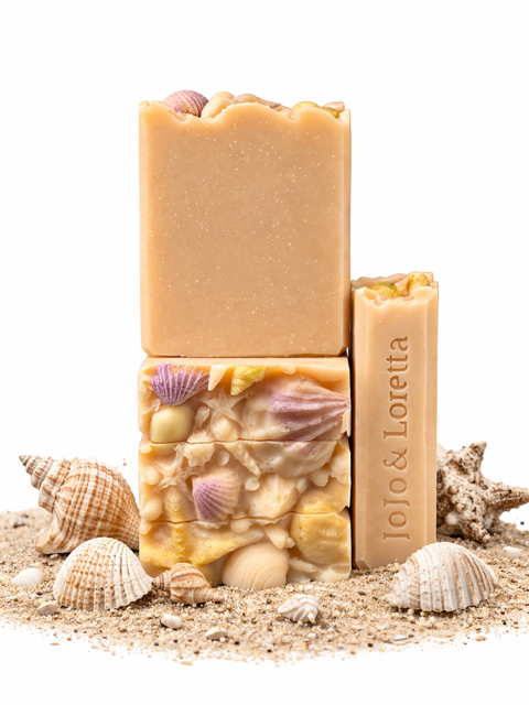 Three bars of soap with shell and starfish decorations on a sandy surface, with additional shells around them.