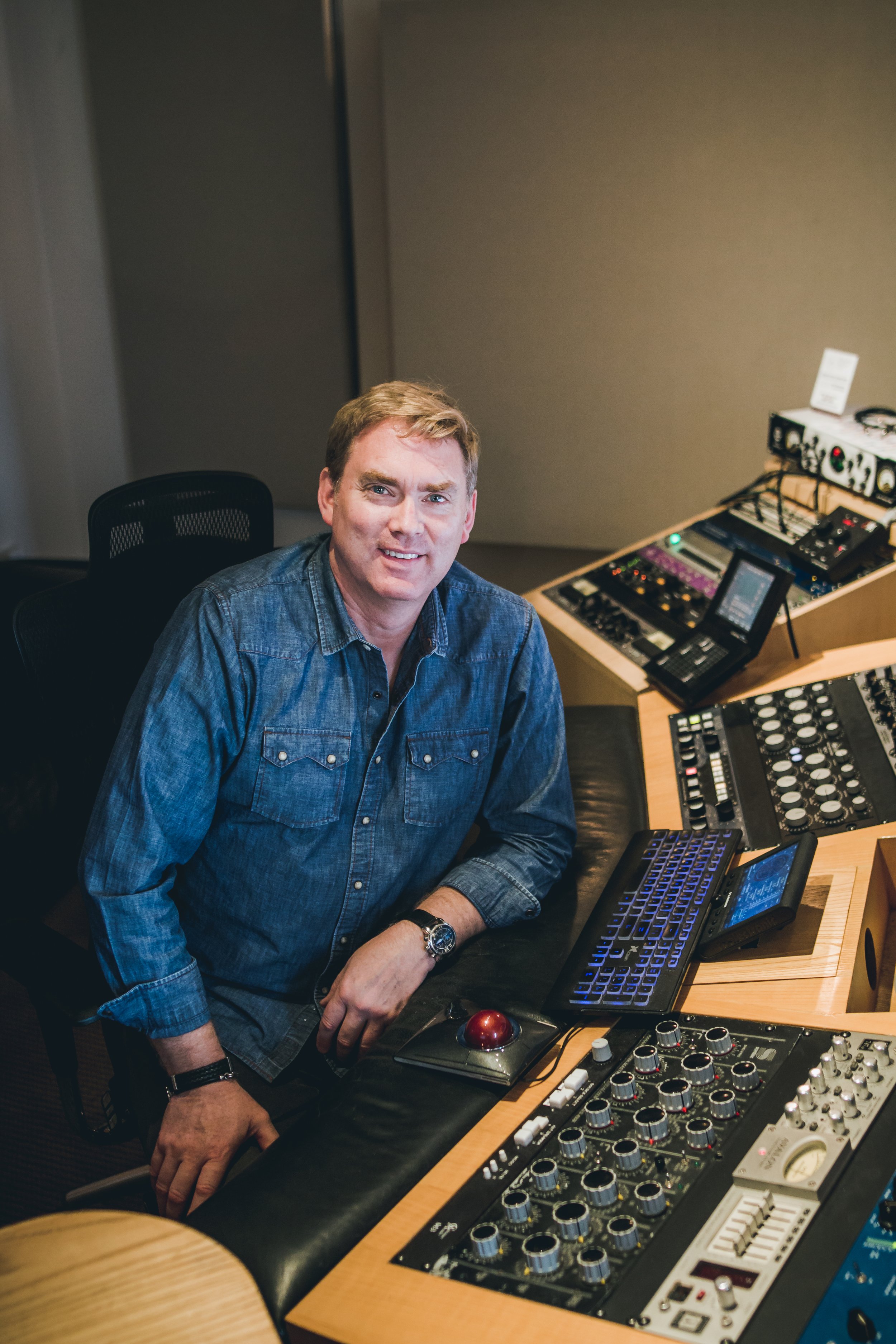 A man in a denim shirt sitting at a recording studio control board.