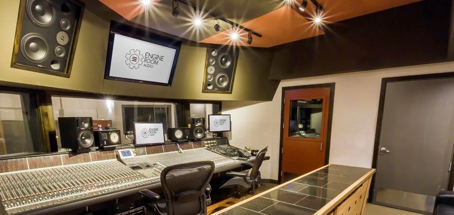 Sound mixing studio with a large mixing console, multiple speakers, two monitors displaying 'Engine Room Audio', a control chair, and a door in the background.