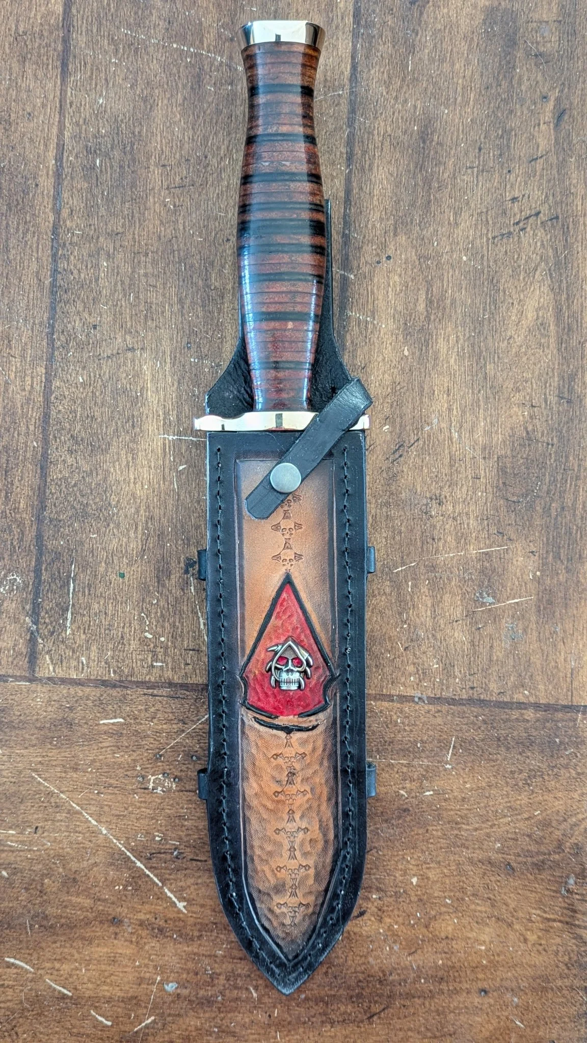 A dagger with a stacked leather handle and a brass guard, in a leather sheath decorated with a skull and dagger design