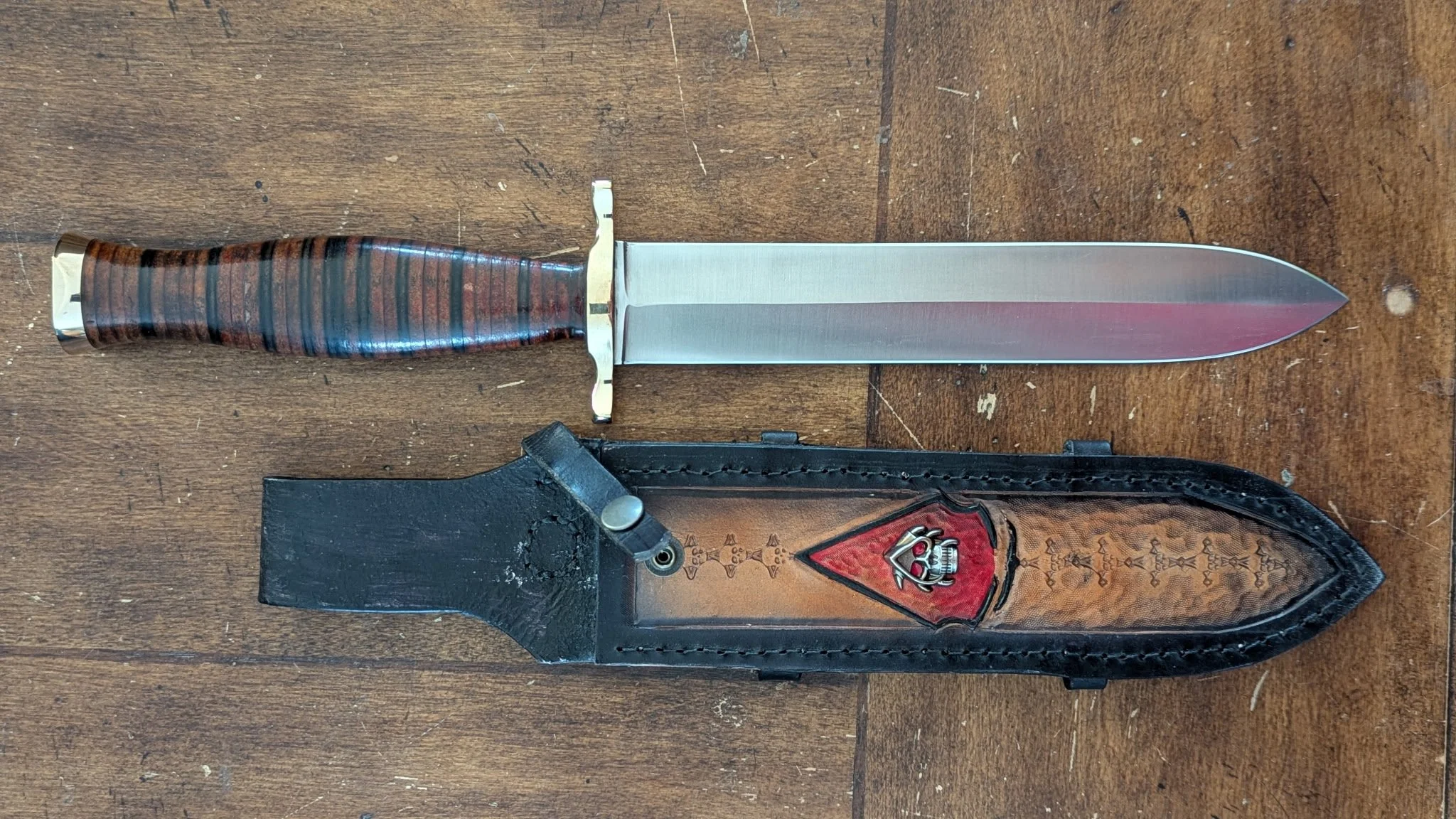 A dagger with a metal blade, stacked leather handle, and a brass guard, placed on a wooden surface. The knife's sheath is black with a red and silver skull emblem and decorative stitching.