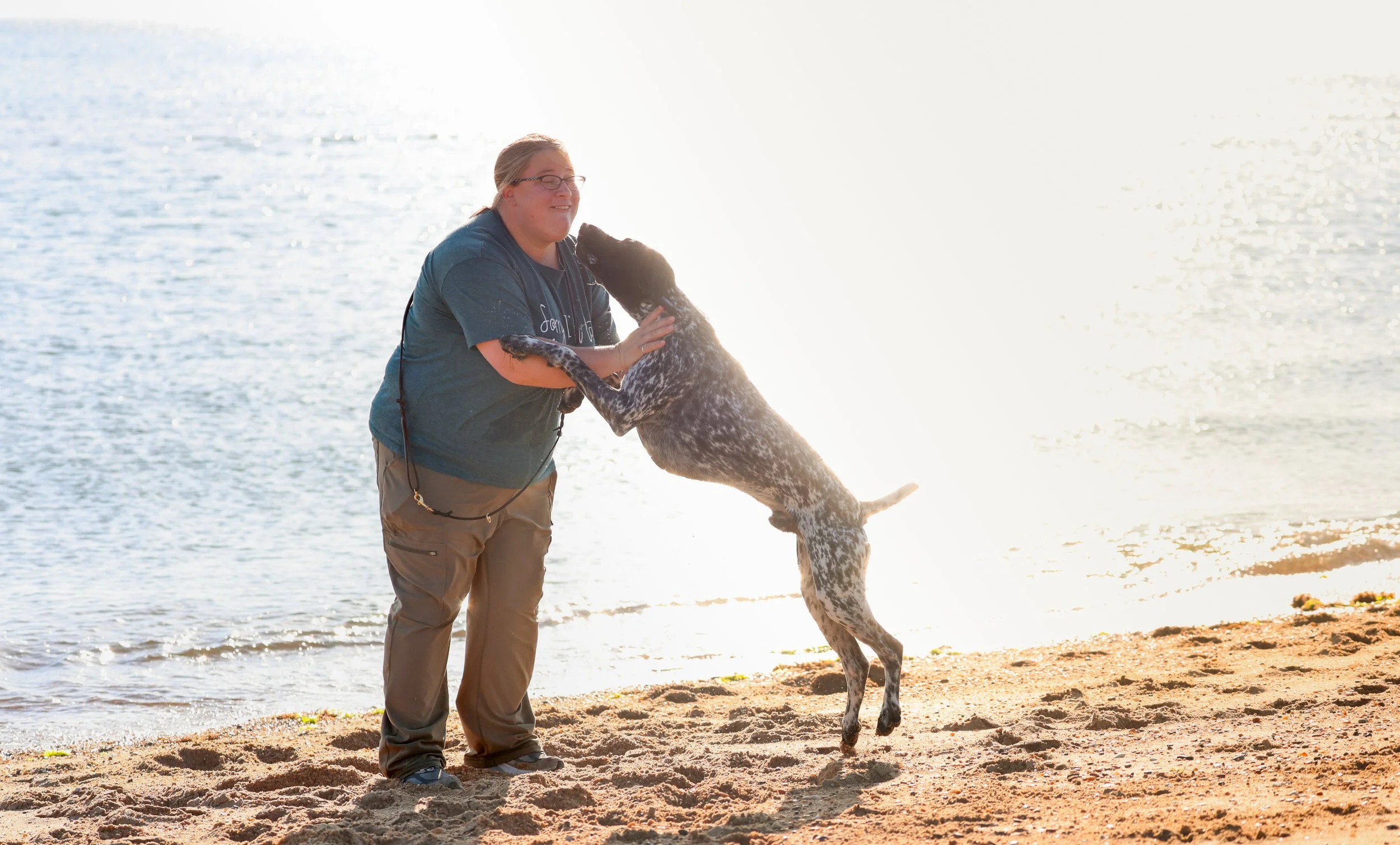 A woman with glasses and a blue t-shirt playing with a large spotted dog on a sandy beach with water and sunlight in the background.