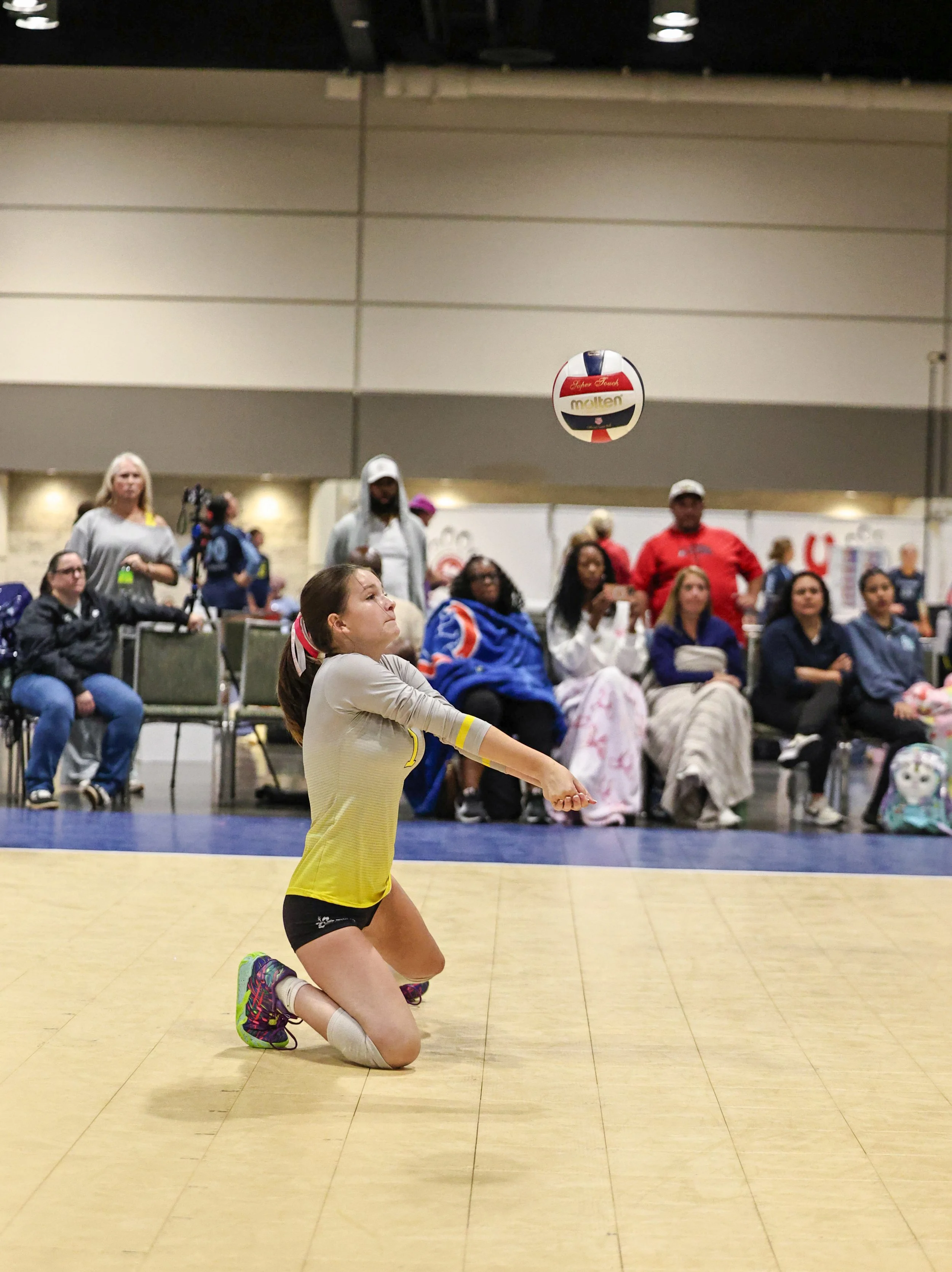 A young girl in a yellow and black sports uniform is kneeling on a volleyball court, with her right knee on the ground and her left leg bent, as she prepares to pass a volleyball. She is wearing colorful athletic shoes. In the background, there is an audience seated and standing, with some people watching the game and others engaging in different activities. The volleyball is in mid-air above her, with the brand name 'molten' visible.
