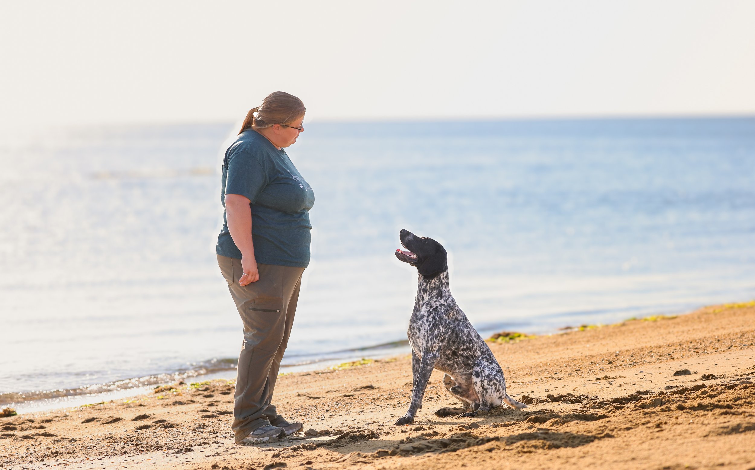 A woman standing on a sandy beach near the ocean, facing a seated black and white spotted dog, both looking at each other.