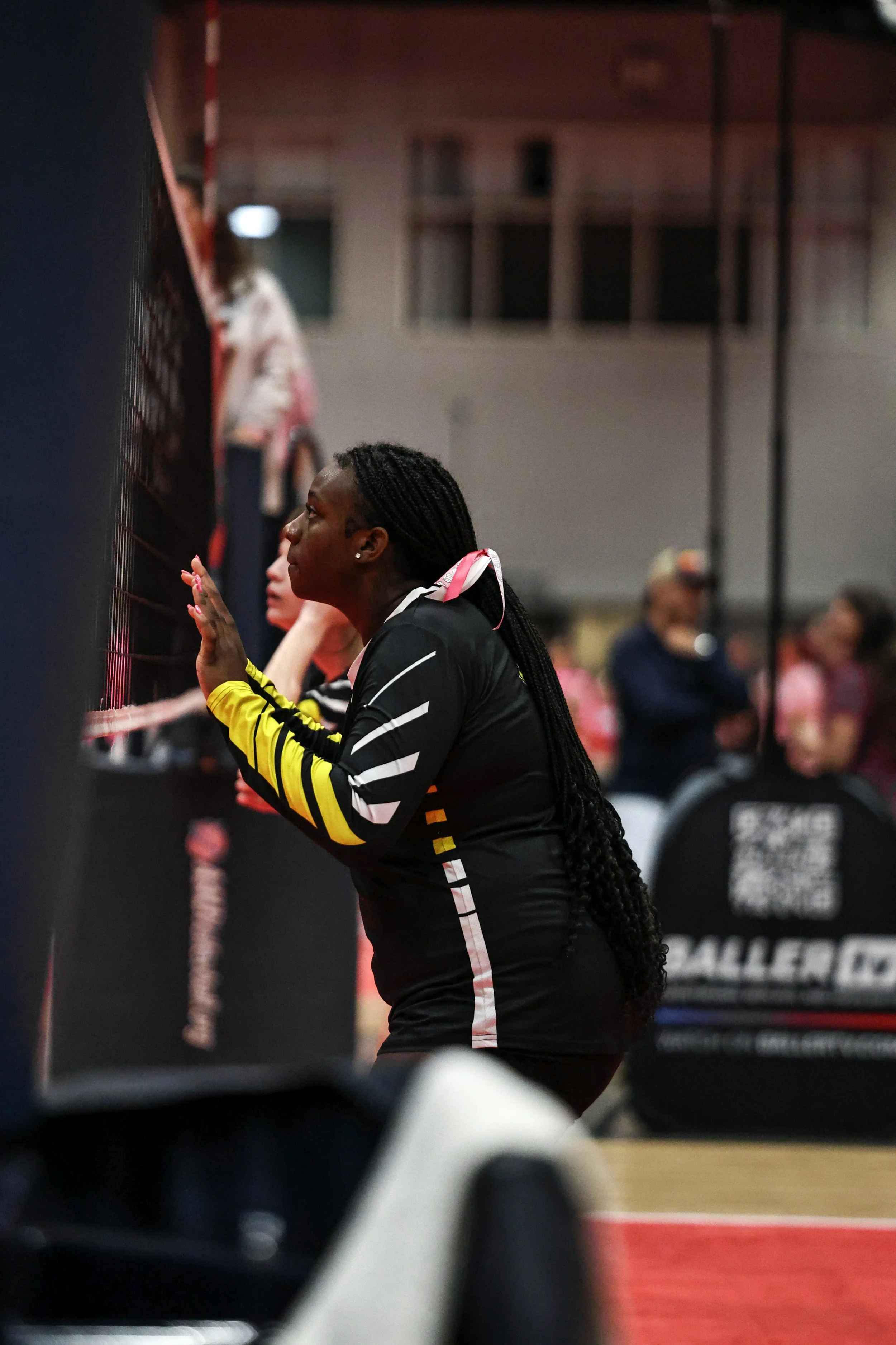 A female volleyball player with long braided hair kneeling on the court, touching the wall. She is wearing a black jersey with pink and white accents, possibly during a game or practice. Other people are blurred in the background, some watching.