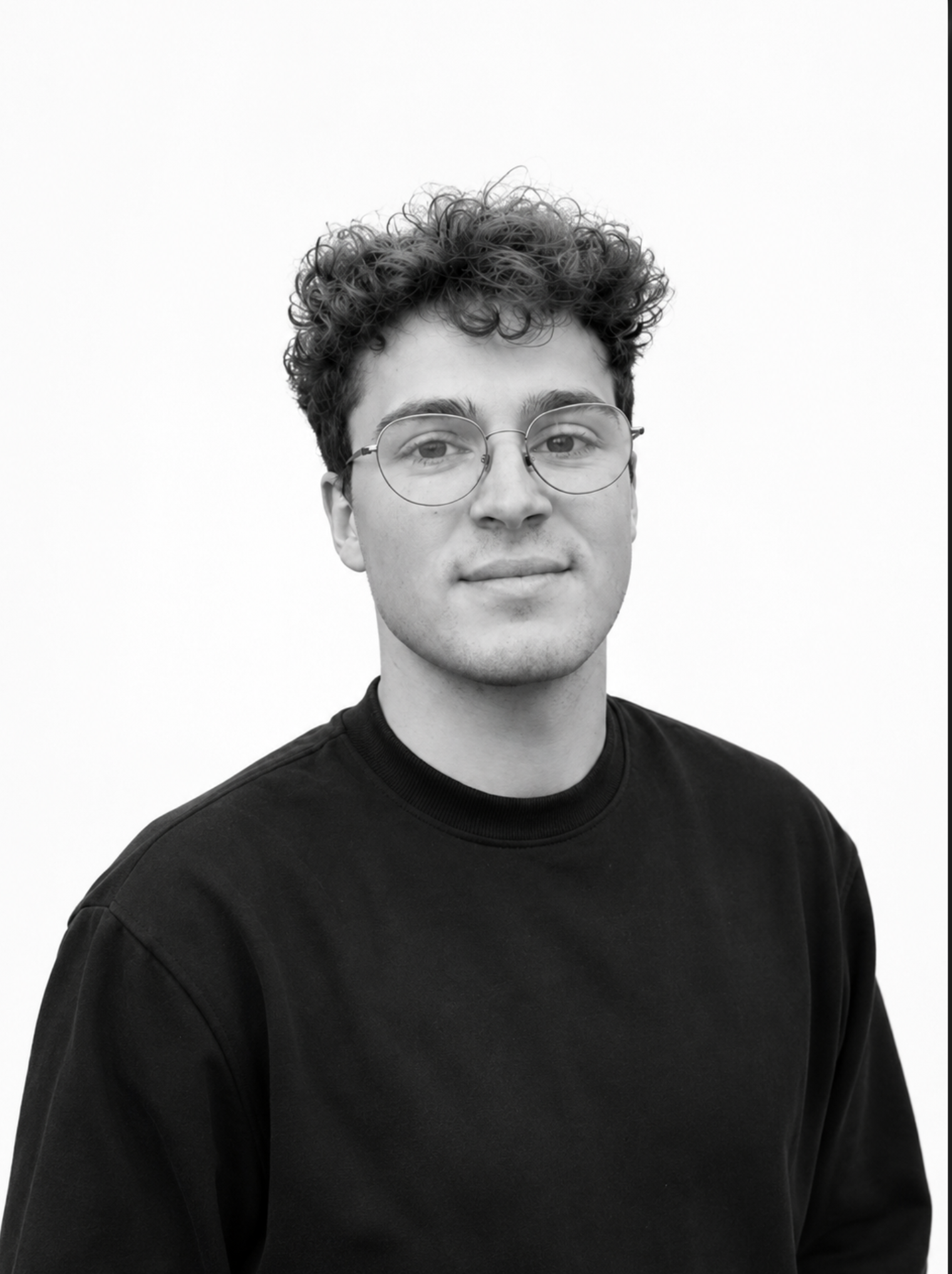 Schwarz-weiße Portraitaufnahme eines jungen Mannes mit Locken und runden Brillengläsern, trägt ein schwarzes Sweatshirt, vor weißem Hintergrund.