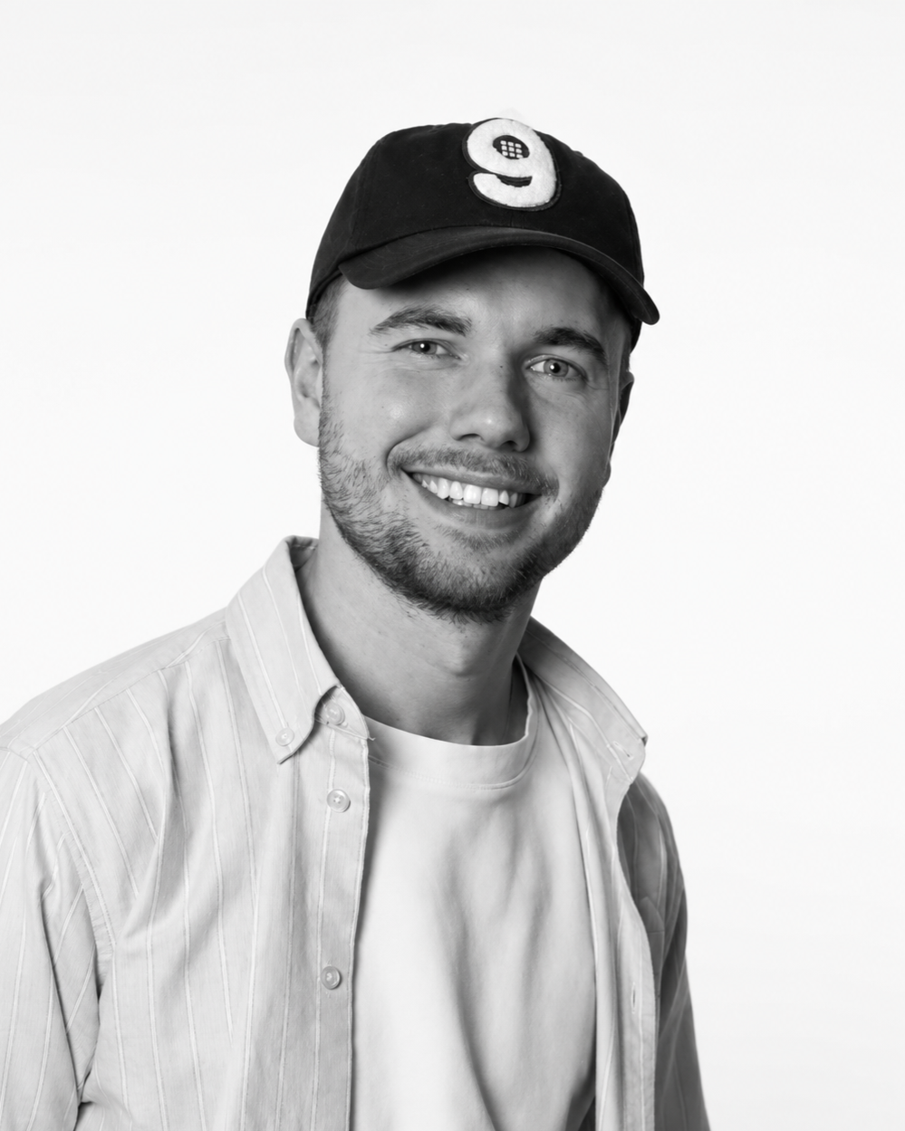 Schwarz-weiß Fotografie eines lächelnden jungen Mannes mit Baseballkappe, hellen Augen, Bart und Backpack, trägt ein Hemd und T-Shirt